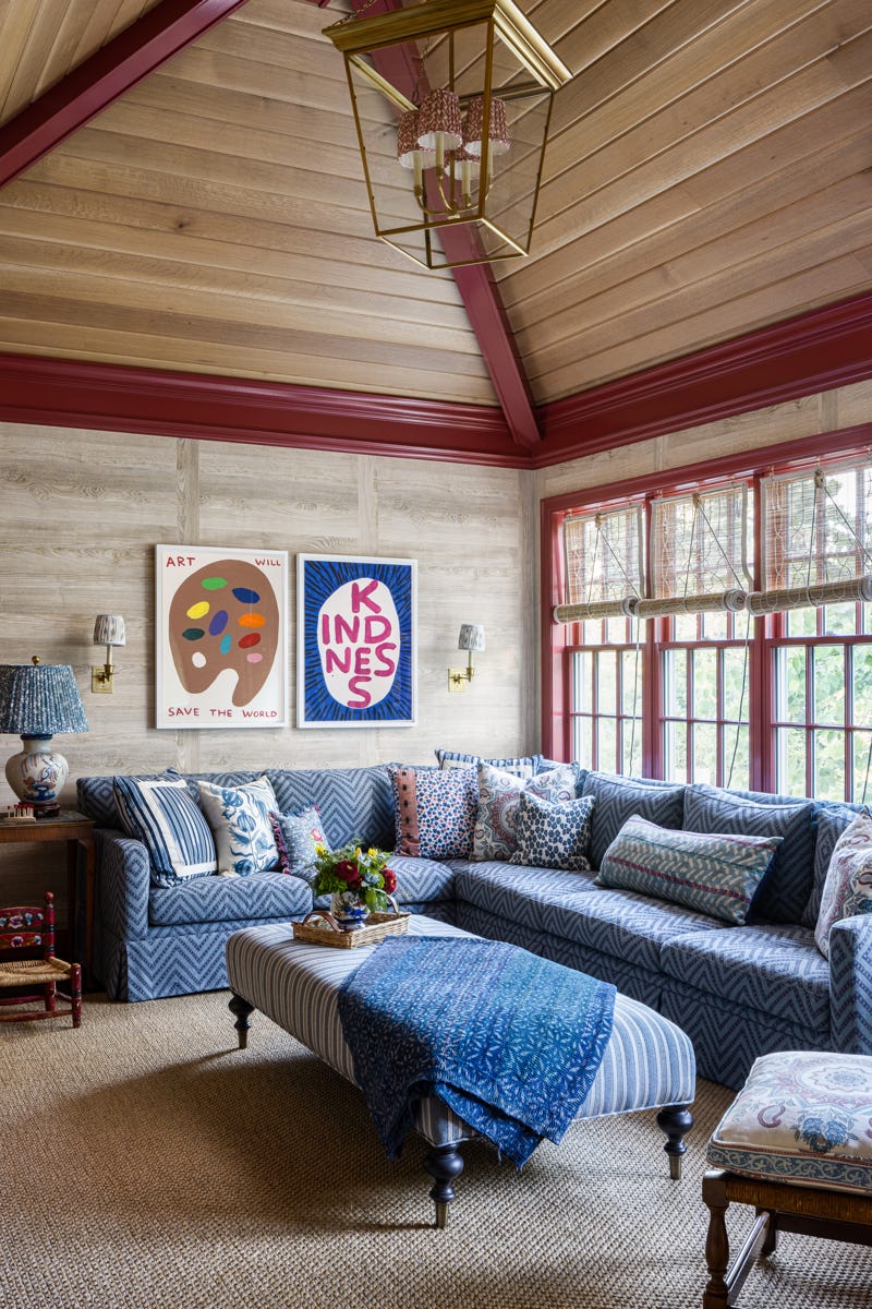 A family room features a blue patterned sectional sofa and striped ottoman under a vaulted wood ceiling with red trim and brass lighting.