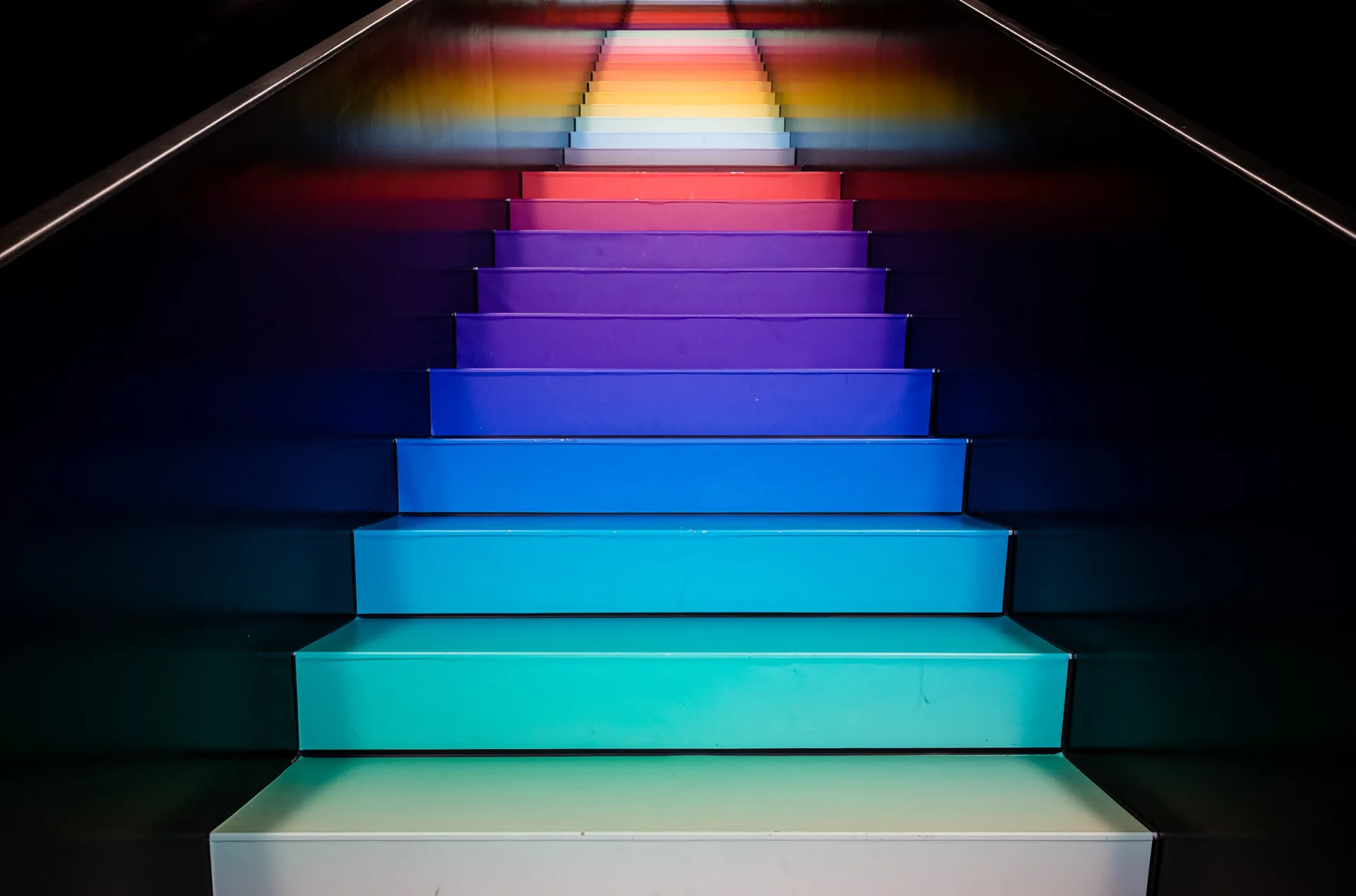 A straight indoor staircase with steps painted in a colorful rainbow gradient, set between two solid matte black walls.