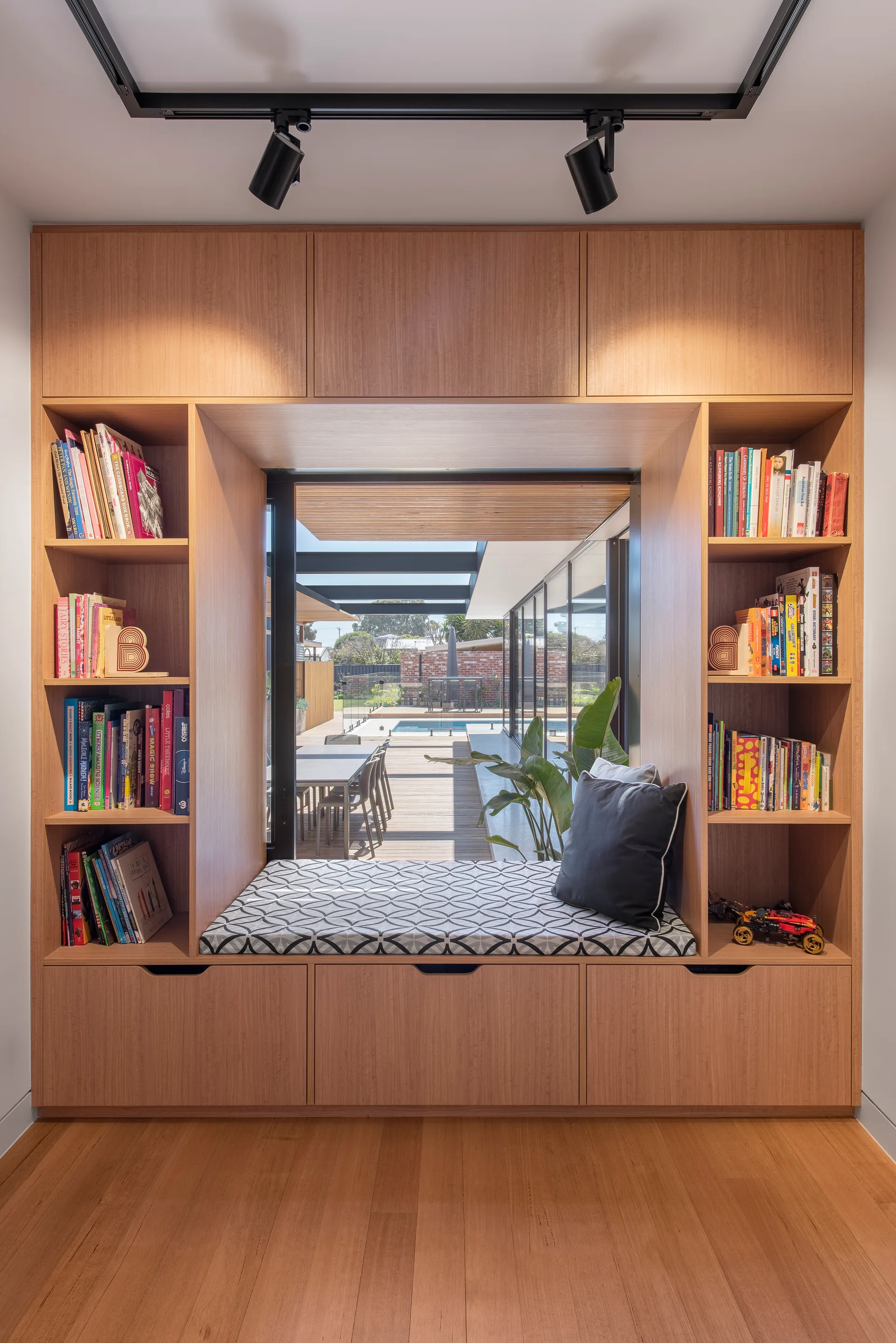 A custom light wood built-in unit features a cushioned window seat flanked by open bookshelves filled with colorful books.