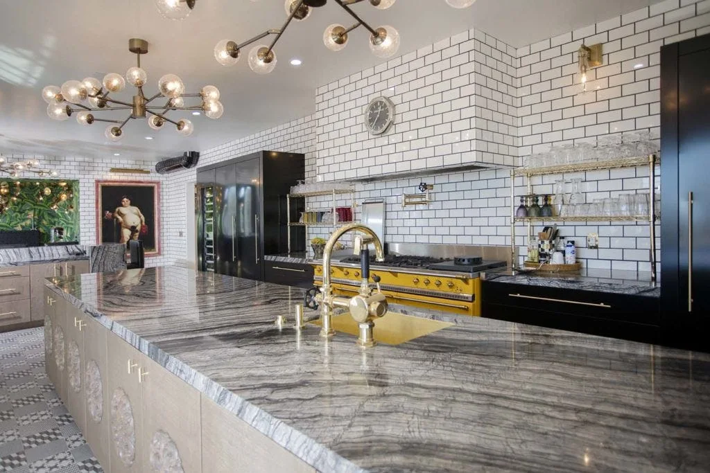 A kitchen features a highly veined grey stone island, a yellow gas range stove, and a white subway tile backsplash.