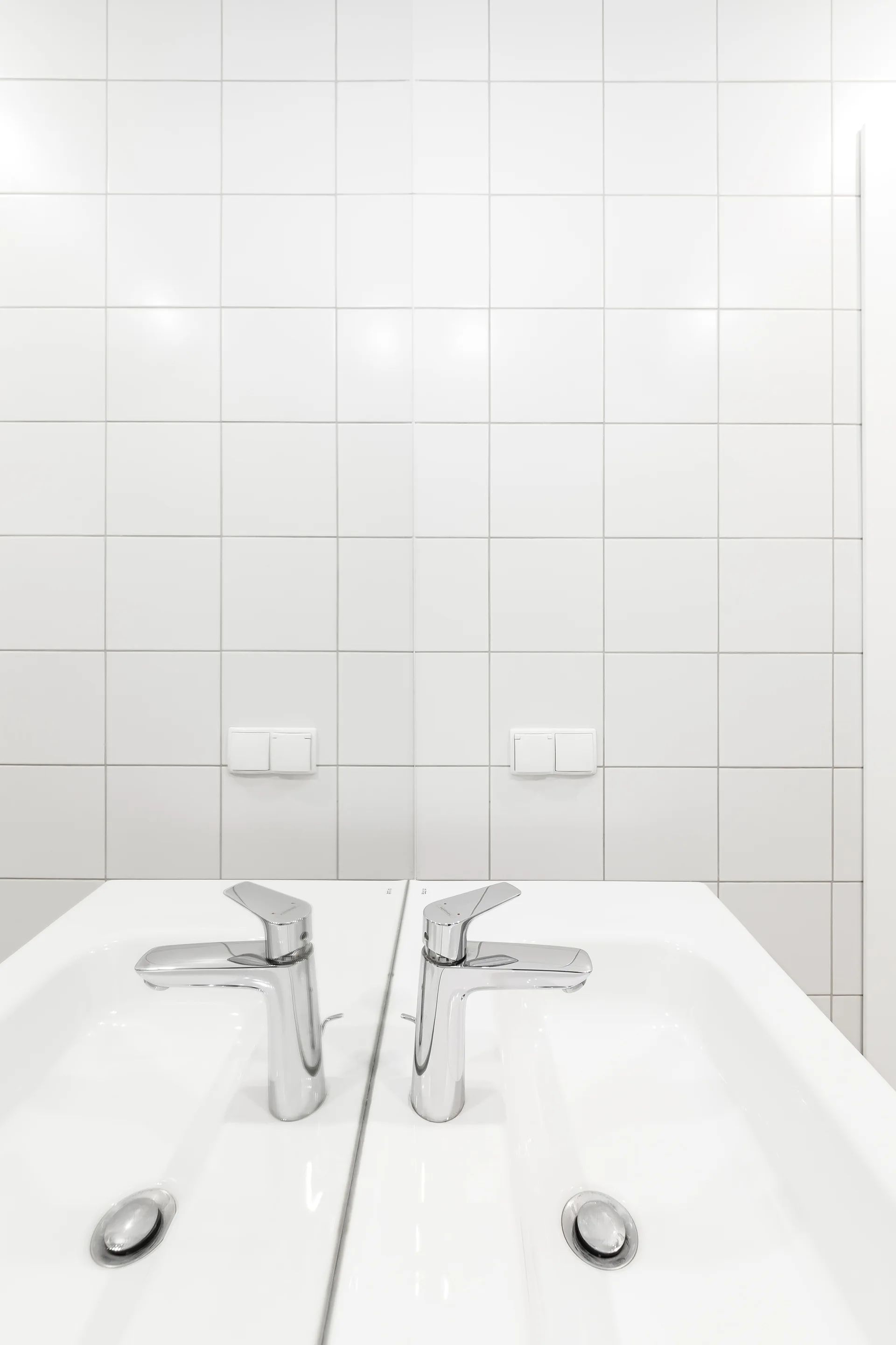 A white ceramic bathroom basin and chrome faucet are reflected in a mirror against a wall of square white grid tiles.