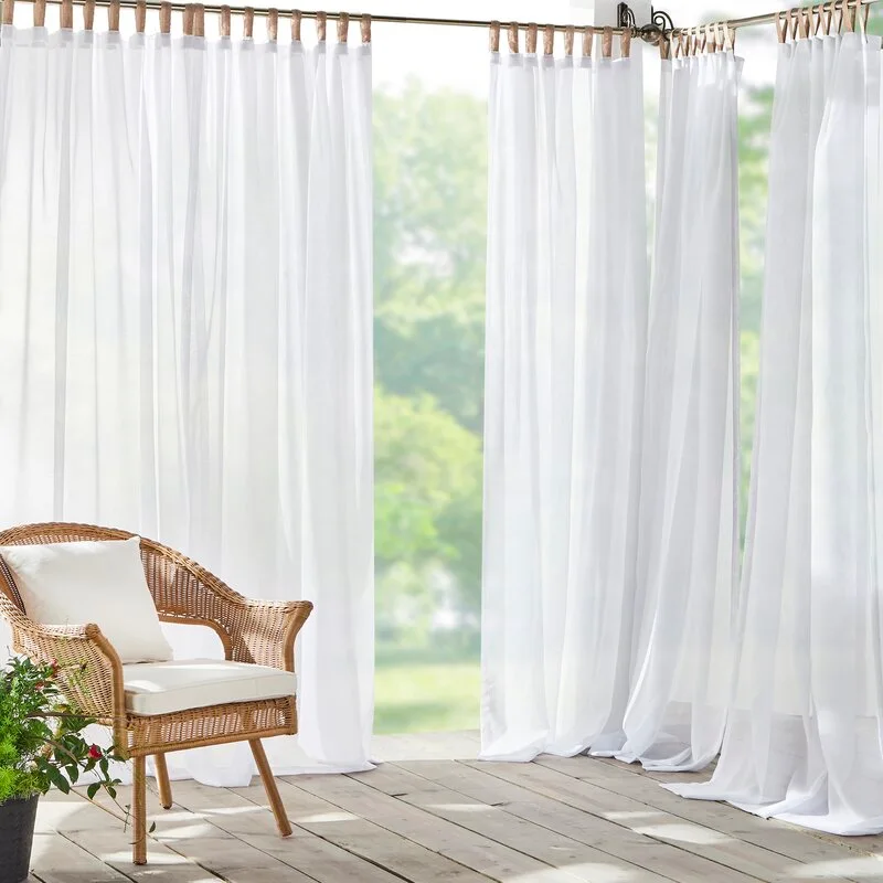 A wicker armchair with white cushions sits on a wooden deck surrounded by flowing, sheer white outdoor curtains under soft natural light.