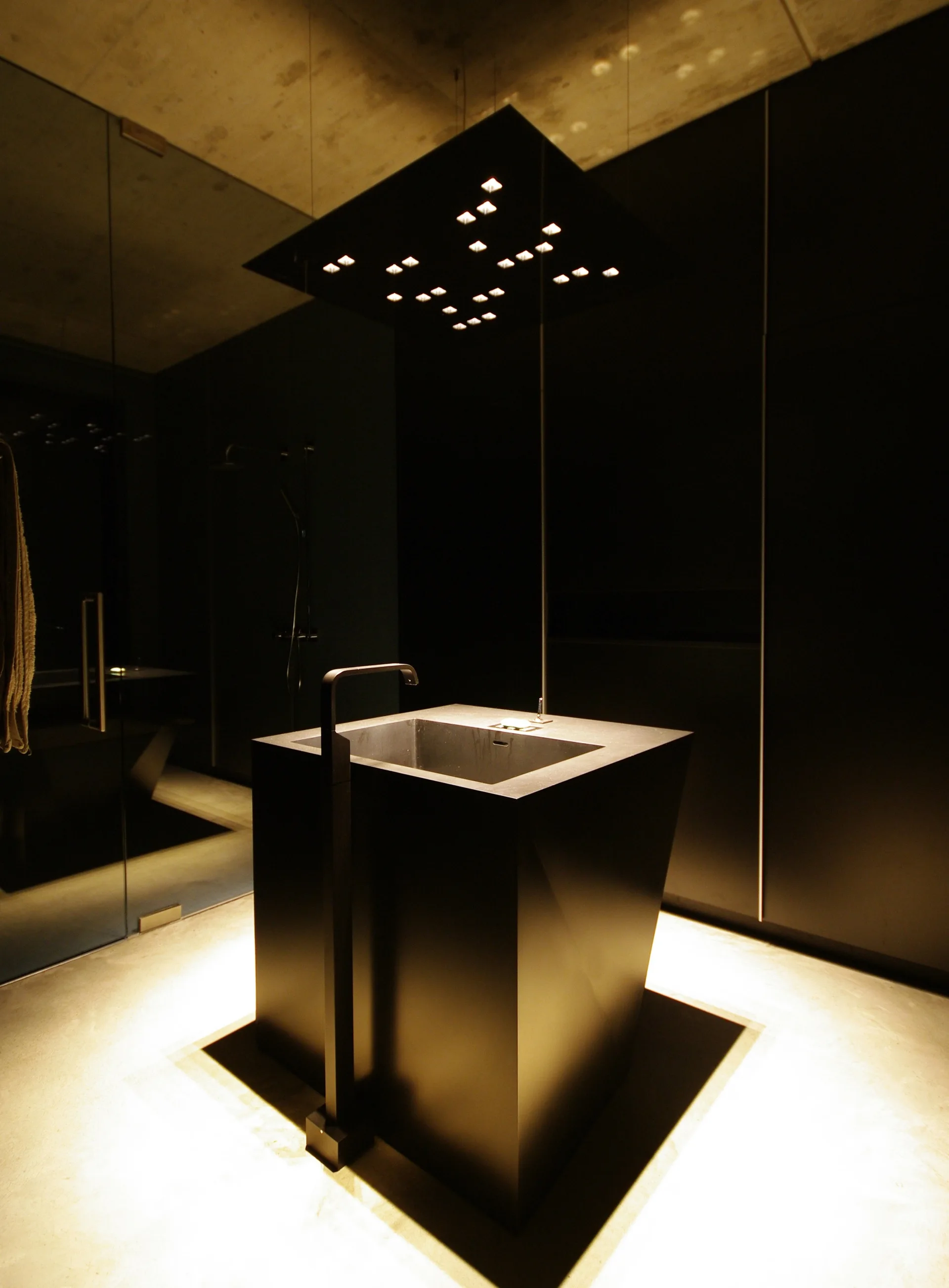 A modern bathroom showcases a monolithic black pedestal basin with a tall floor-mounted faucet beneath a square ceiling spotlight array.