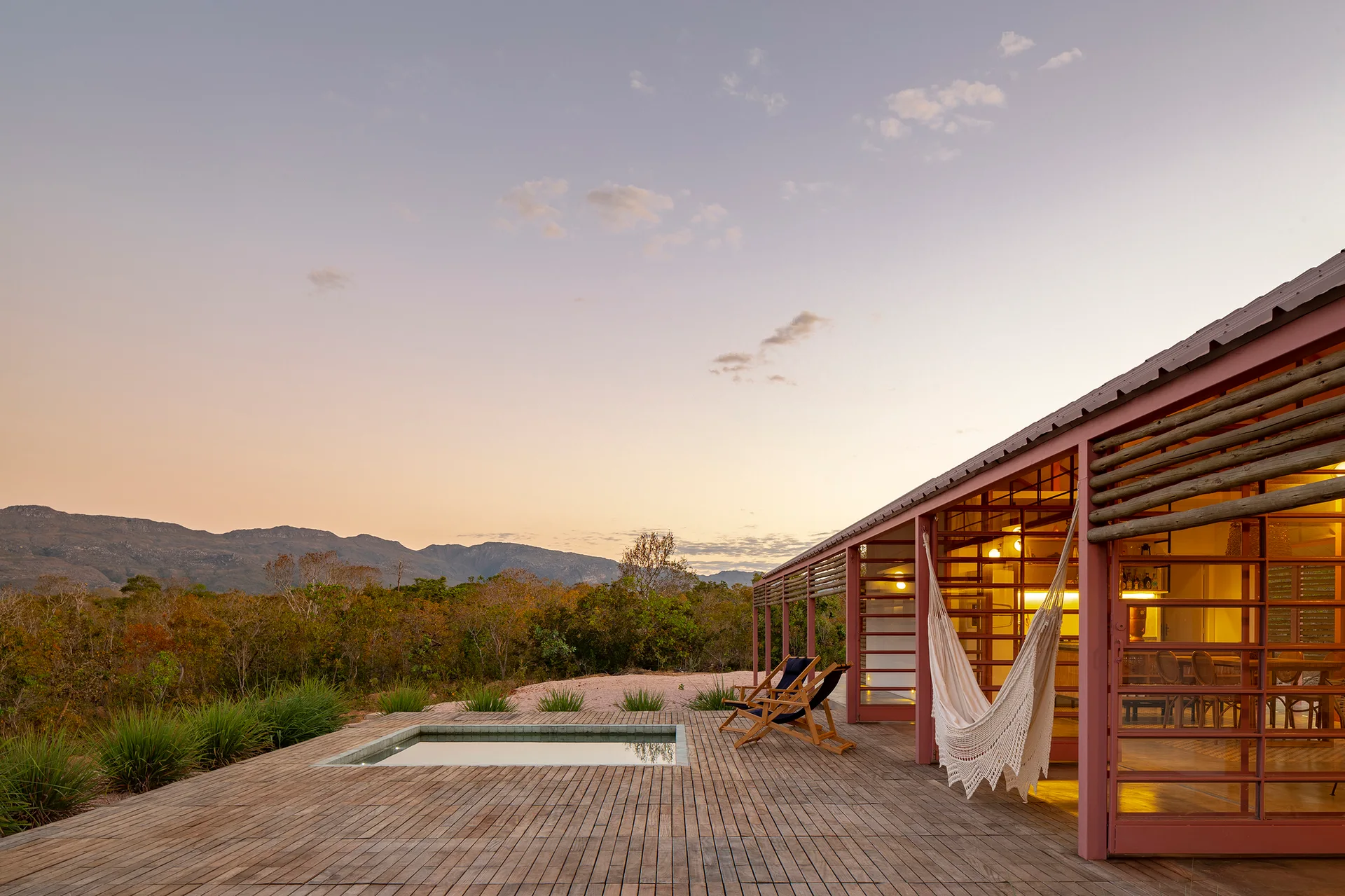 A wide wooden deck features an embedded square plunge pool, a white knitted hammock, and lounge chairs against a sunset sky and mountain landscape.
