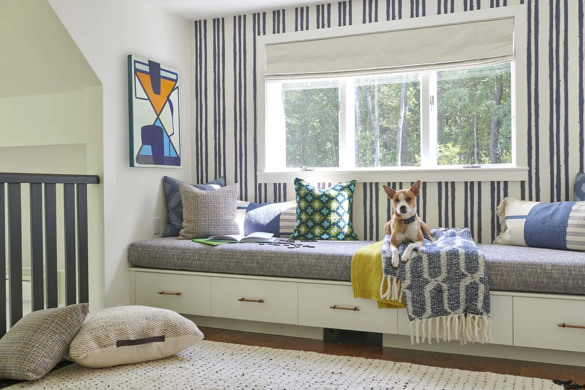 A built-in white storage bench with a grey cushion and colorful pillows is set against a blue and white striped wall beneath a window.