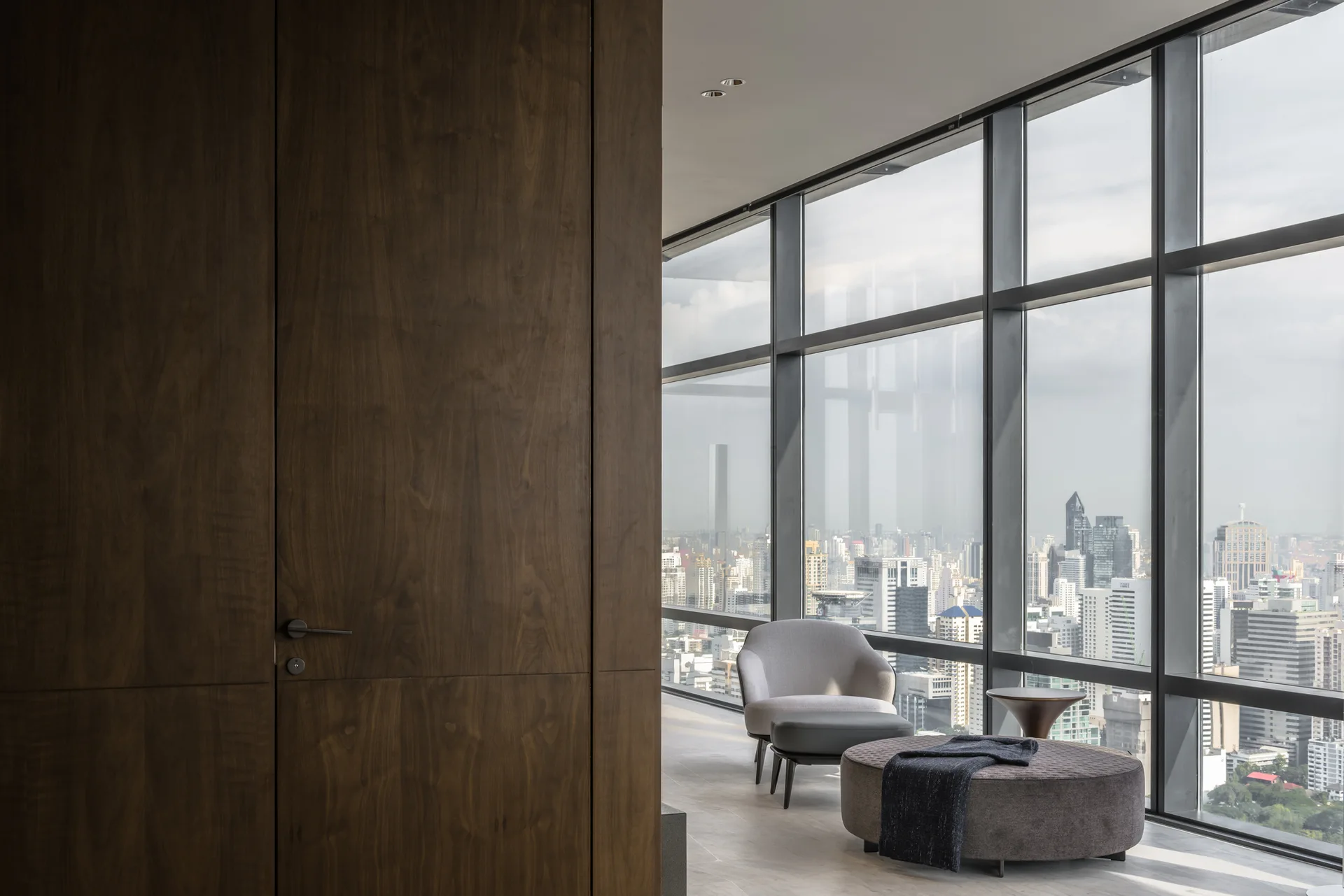 A minimalist gray armchair and round ottoman sit near large windows overlooking a city skyline, next to a tall walnut wood paneled wall.