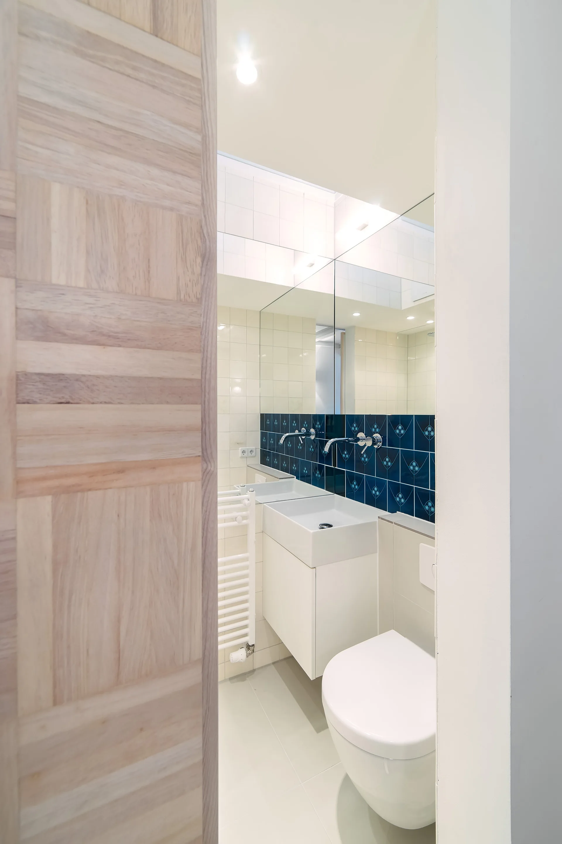 View through a wooden doorway of a compact white bathroom with a wall-mounted sink, toilet, large mirror, and blue patterned tile backsplash.