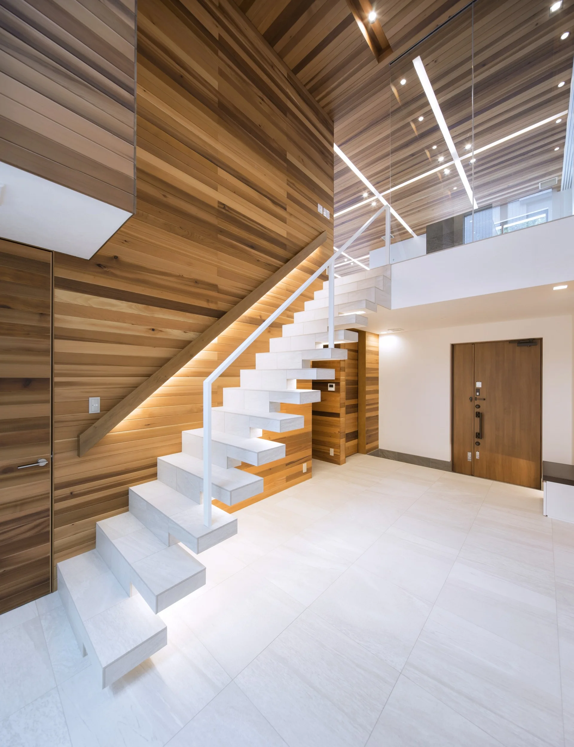 A modern foyer features a white floating stone staircase set against warm horizontal wood-paneled walls and a wood plank ceiling.