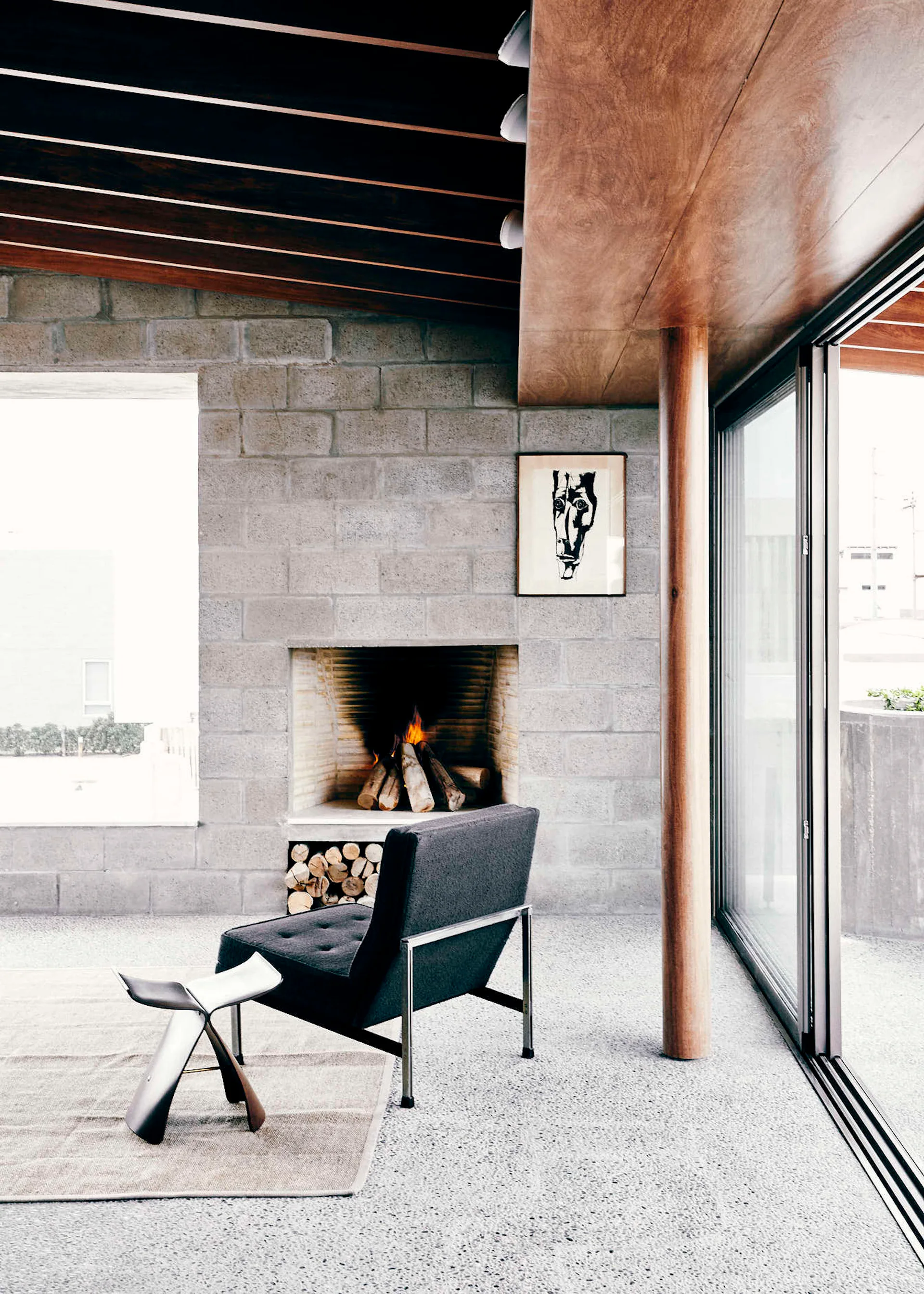 A modern living room features a concrete block fireplace, a dark grey armchair, a wooden support column, and a slatted ceiling.