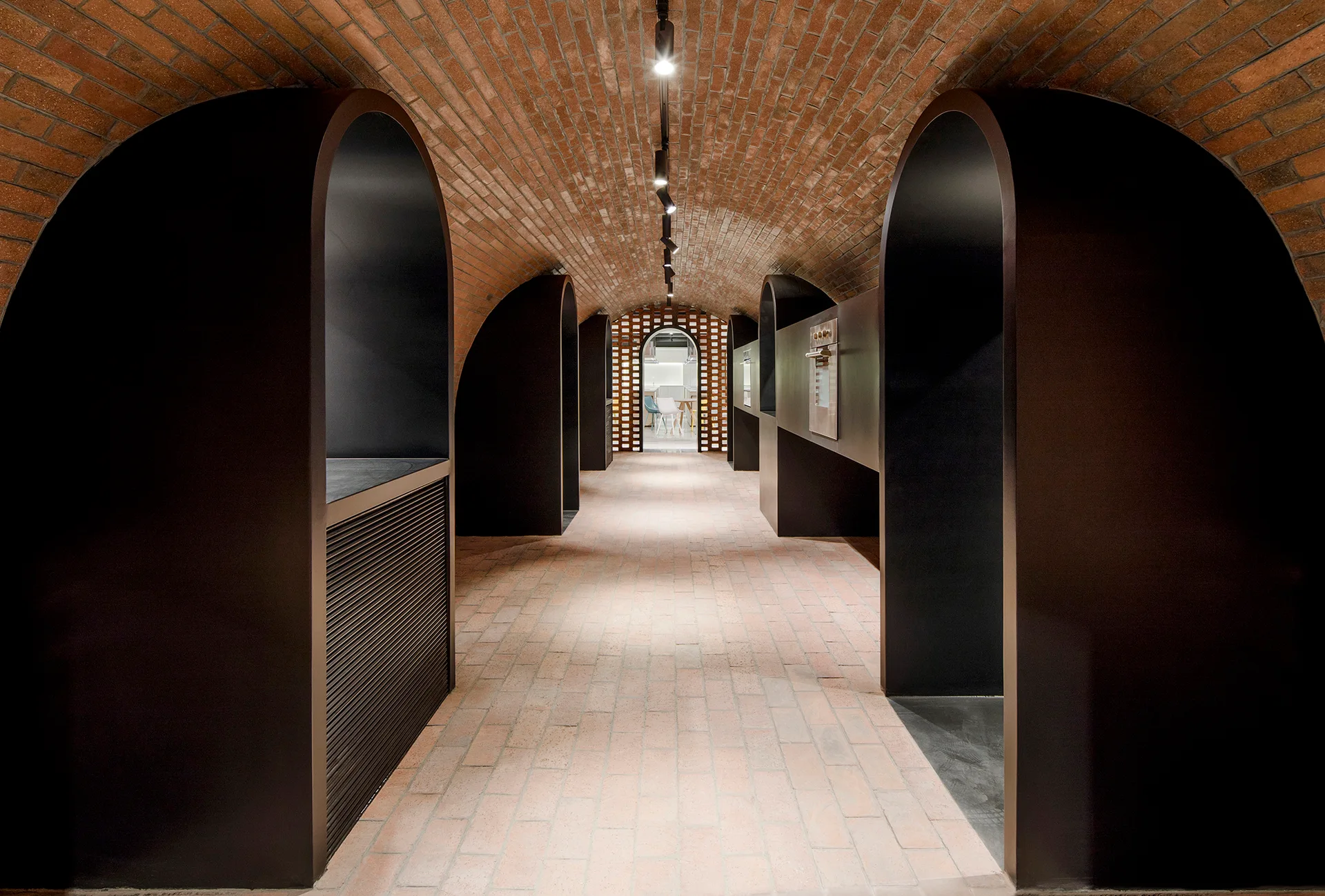 A long hallway with a curved red brick barrel vault ceiling and floor features repeating matte black arched wall partitions and track lighting.