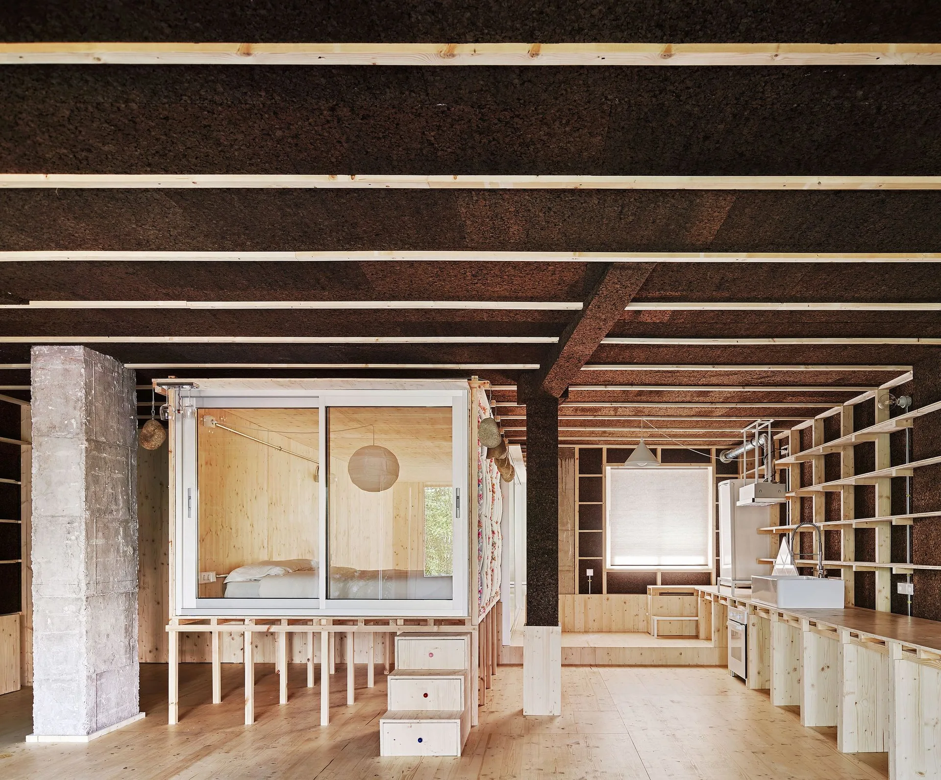 An open loft features a glass-enclosed sleeping platform, minimalist light timber kitchen counters, and a dark textured cork ceiling.