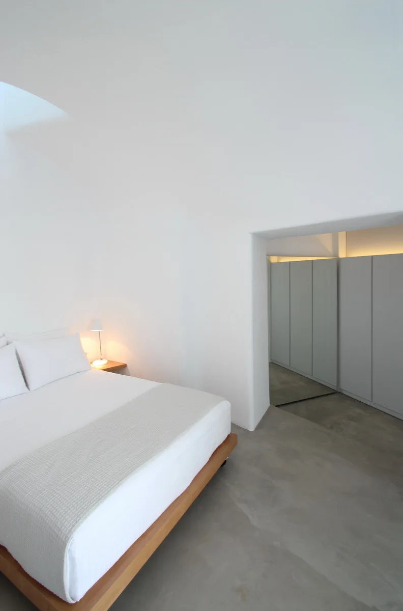 A minimalist white bedroom features a wooden platform bed on concrete floors under a vaulted ceiling, with a wardrobe alcove visible in the background.