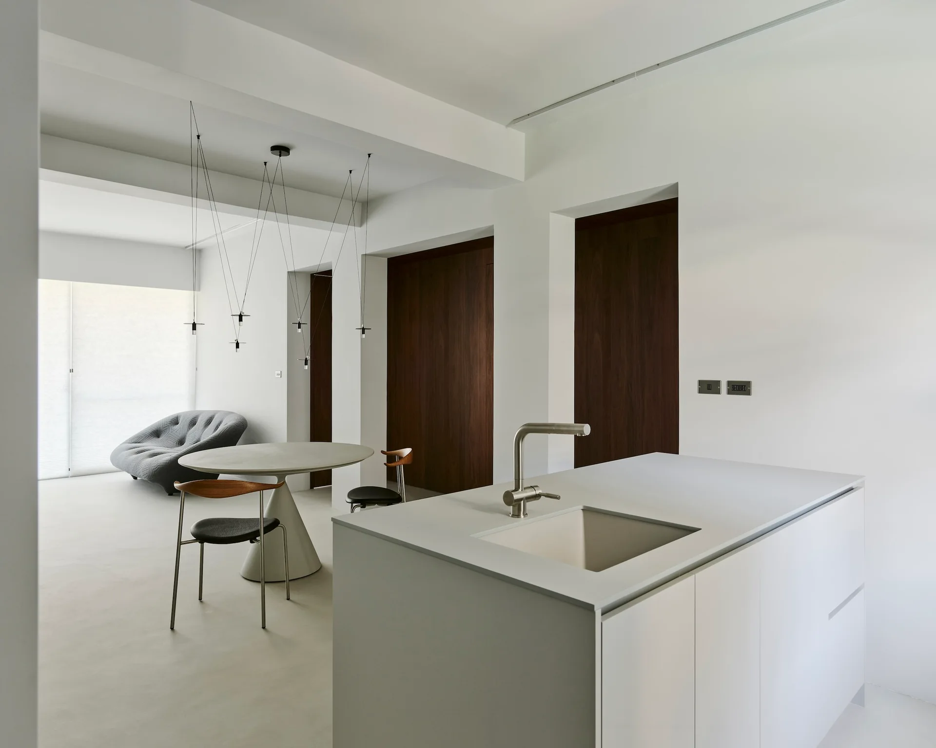 A minimalist open-plan kitchen featuring a grey island with an integrated sink, a round pedestal dining table, and dark wood doors.