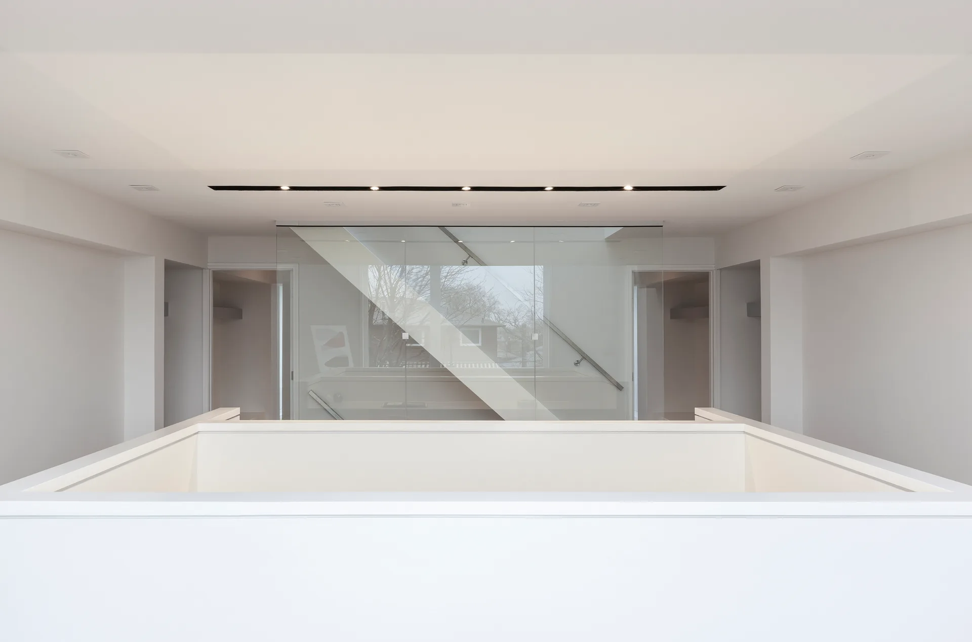 A minimalist white hallway landing with a solid lower railing and a central floor-to-ceiling glass partition showing a staircase.