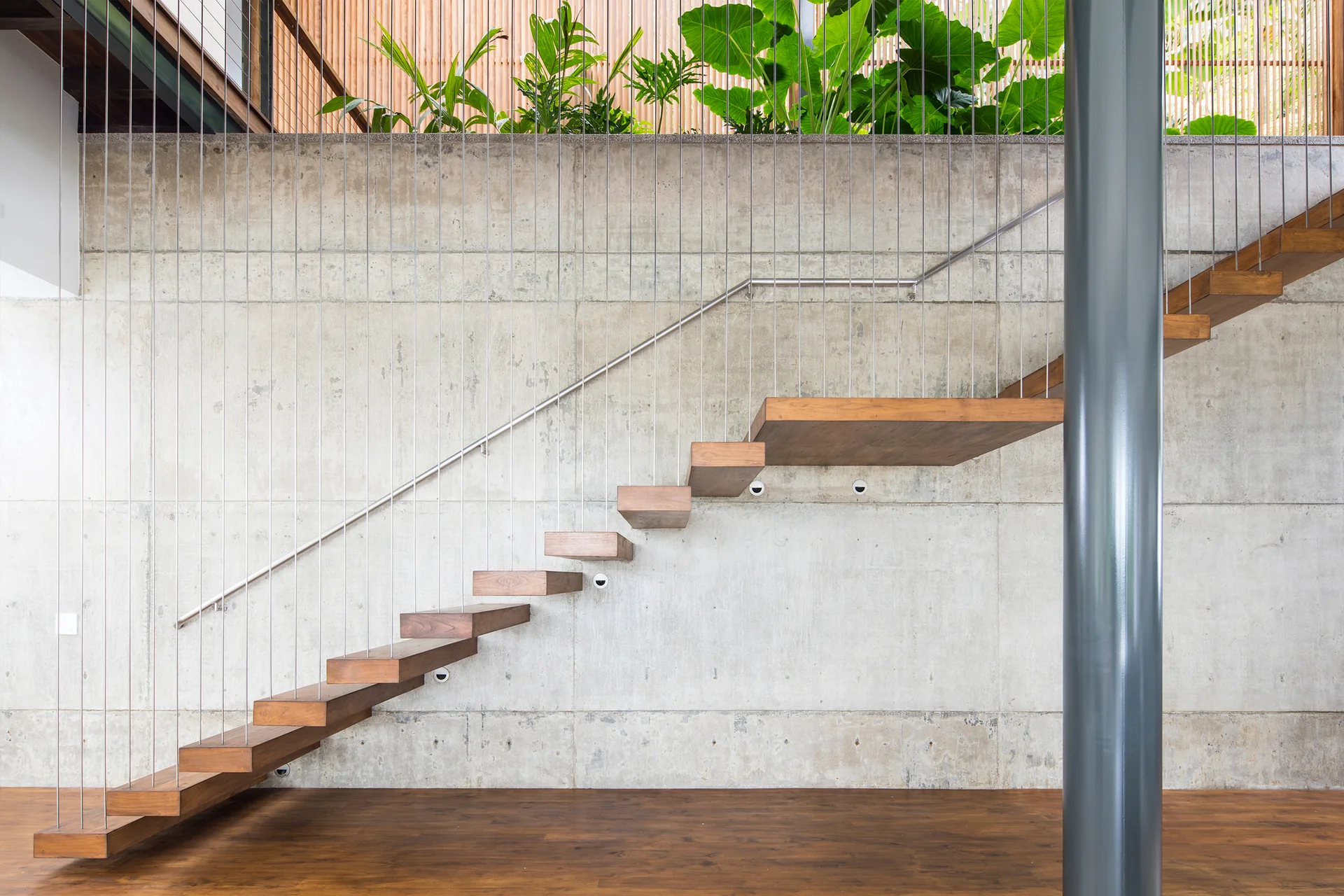 A floating wooden staircase with thin vertical cable railings is mounted against a textured grey concrete wall next to a grey pillar.