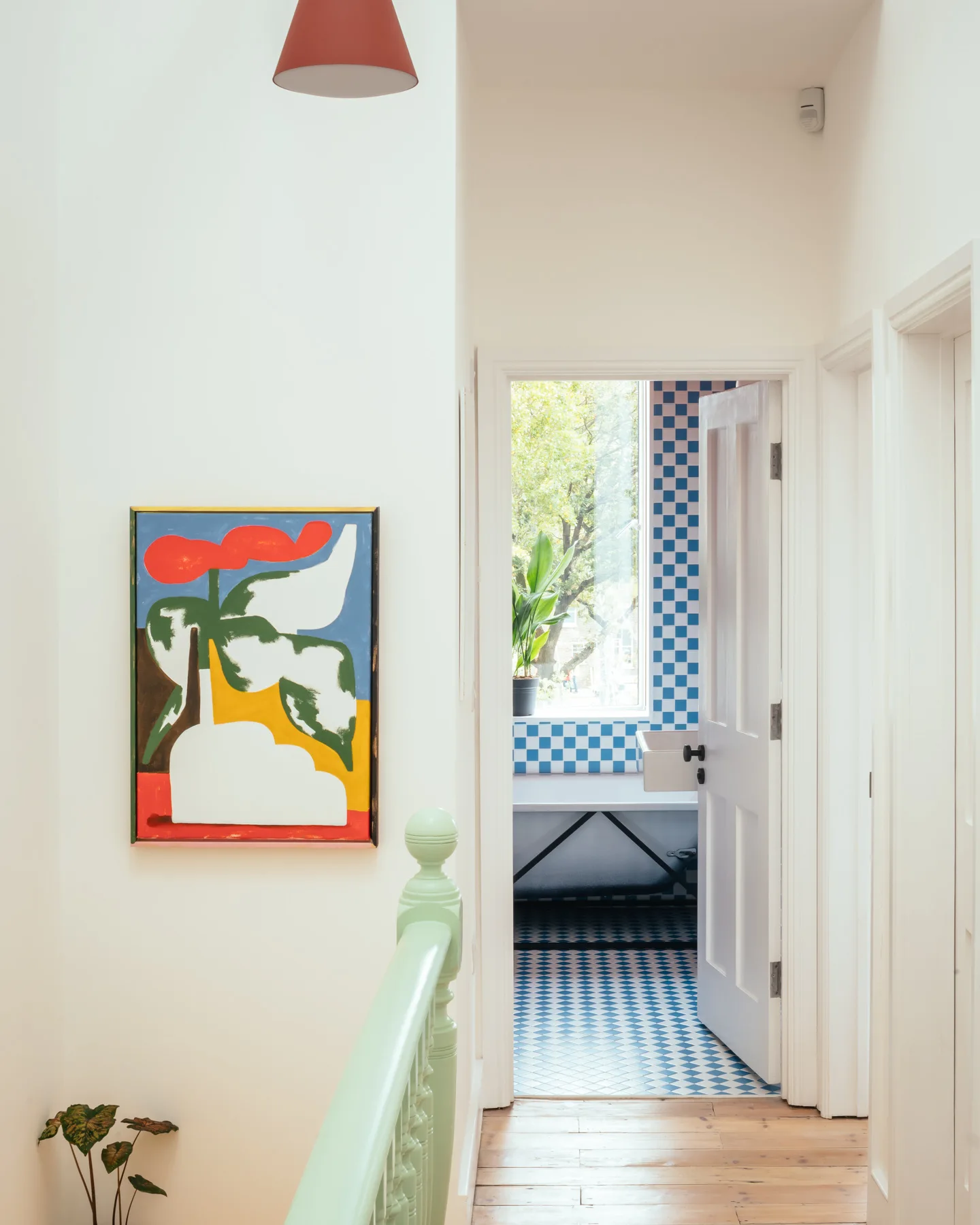 A white hallway with a light green banister and colorful art leads to an open bathroom featuring blue and white checkerboard floor and wall tiles.