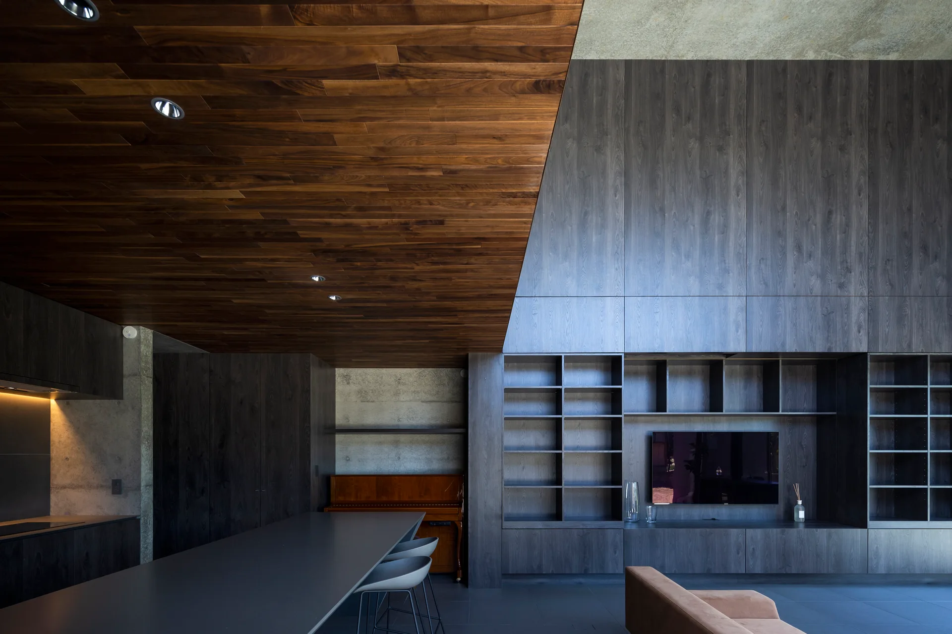 A modern open-plan living room featuring a sloped wood plank ceiling, integrated charcoal wood wall shelving with a television, and concrete walls.