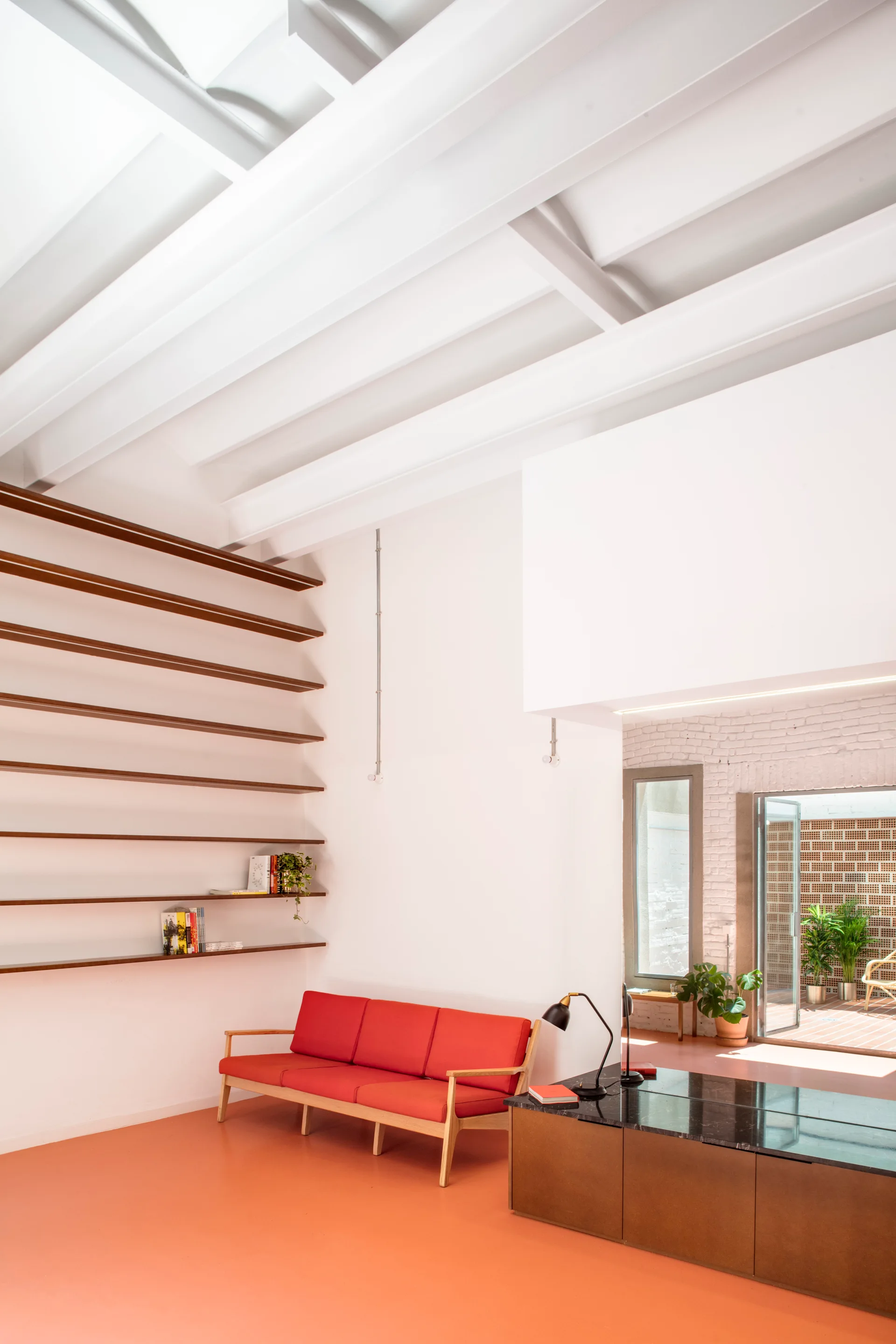 A minimalist living room featuring a bright red sofa, a coral orange floor, long floating shelves on a white wall, and exposed white ceiling beams.