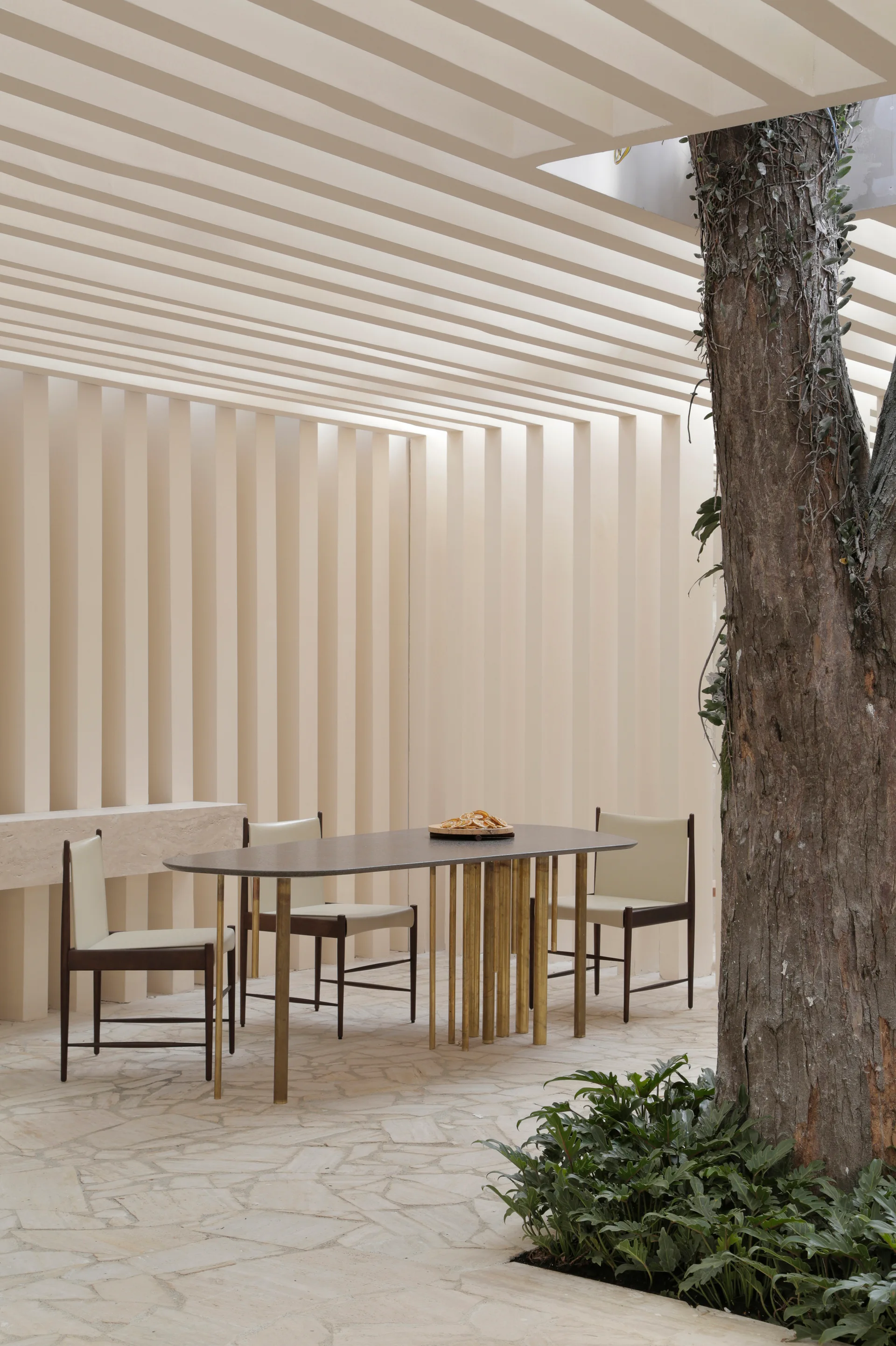 A modern cream patio features an oval dining table under a slatted ceiling, positioned near a large tree trunk and a vertical slatted wall.