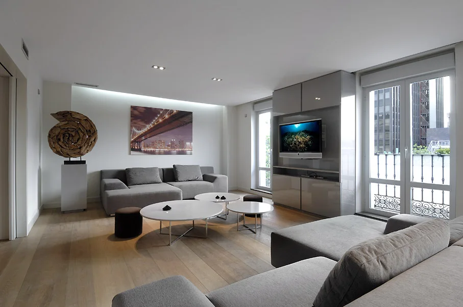 A contemporary living room features a grey sectional sofa, white nested coffee tables, a glossy grey media cabinet, and light wood flooring.