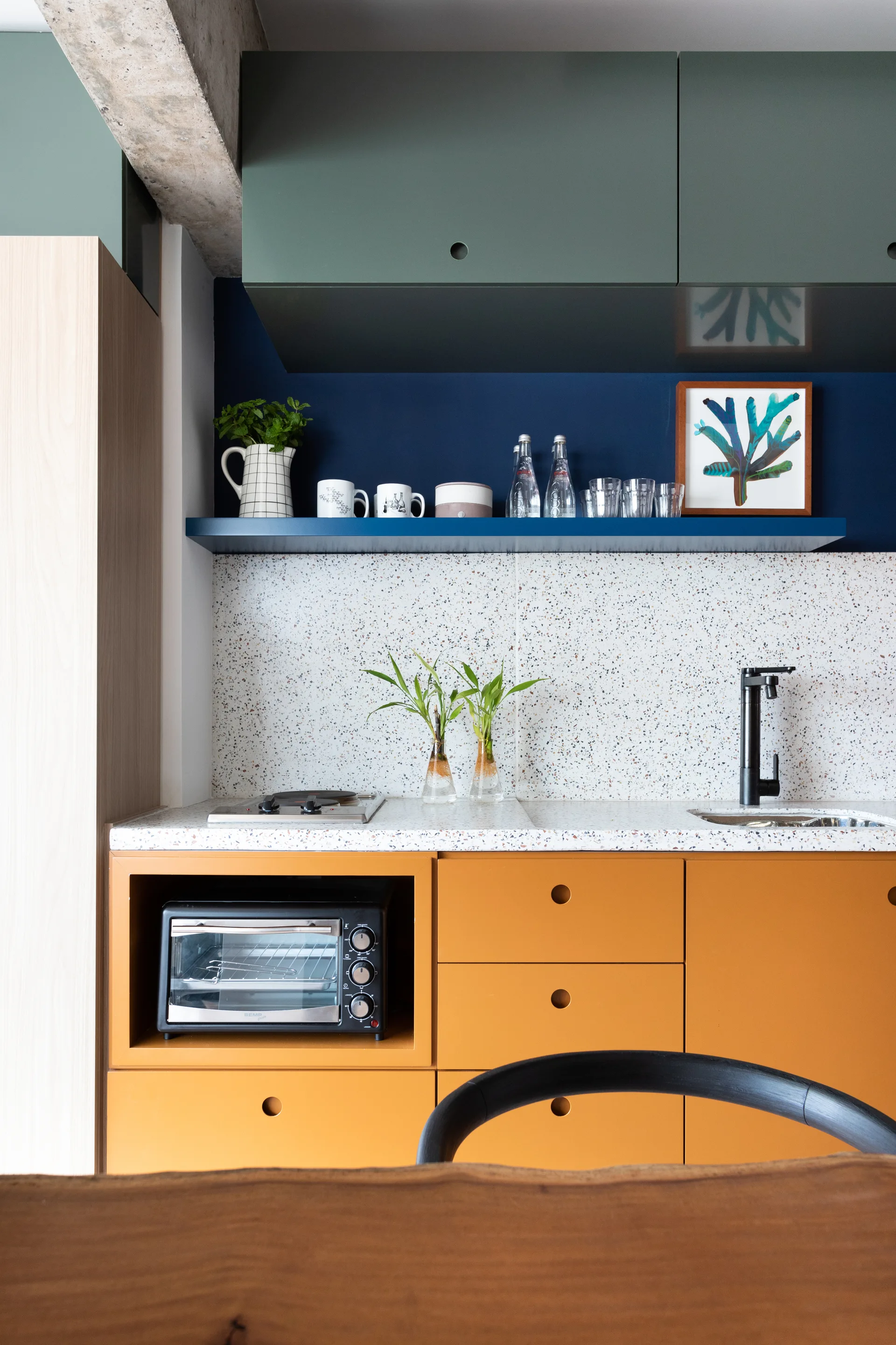 A kitchen features orange base cabinets, a terrazzo countertop, green upper cabinets, and a blue wall with a floating shelf.