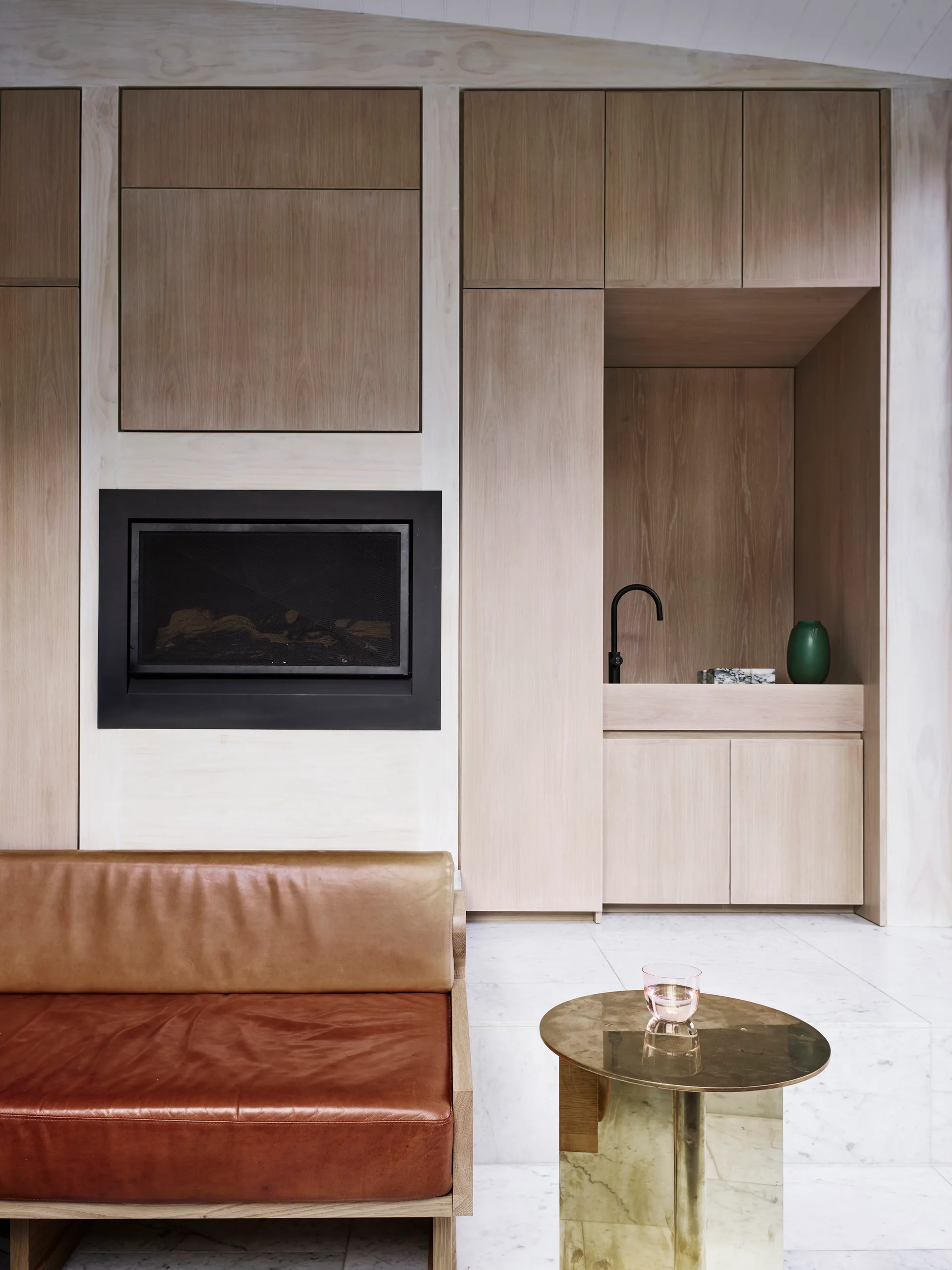 A modern living space features a tan leather sofa, a gold side table, and a wall of light wood built-in cabinetry integrating a fireplace and a wet bar.