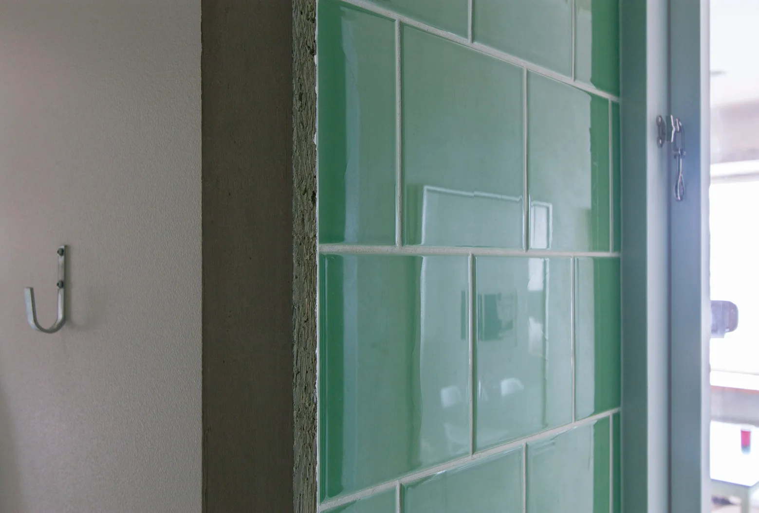 A close-up view of vibrant green glossy ceramic tiles on a wall next to an unfinished edge and a metal hook.