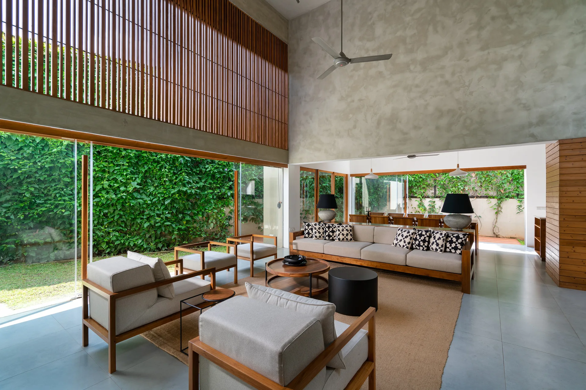 A modern living room features wood-framed sofas on a jute rug beneath high concrete walls and a large glass window looking out onto a lush green garden.