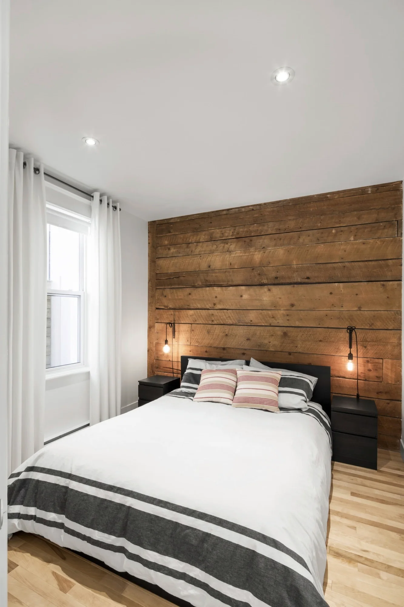 A bedroom with a striped bed set against a horizontal wood plank accent wall, flanked by black nightstands and hanging exposed-bulb sconces.