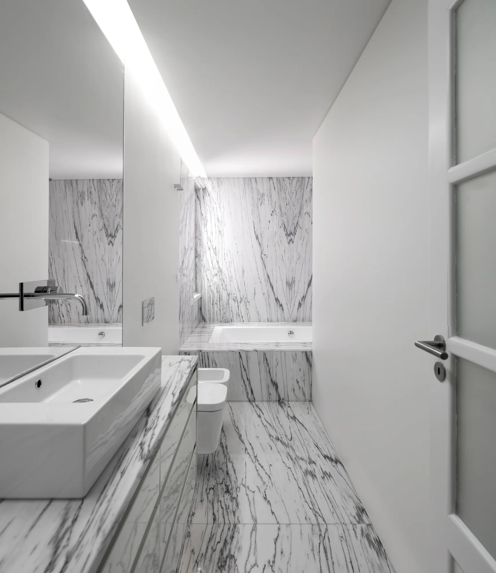 A narrow bathroom features heavily veined white marble on the floors, walls, and vanity base alongside a white vessel sink and built-in bathtub.