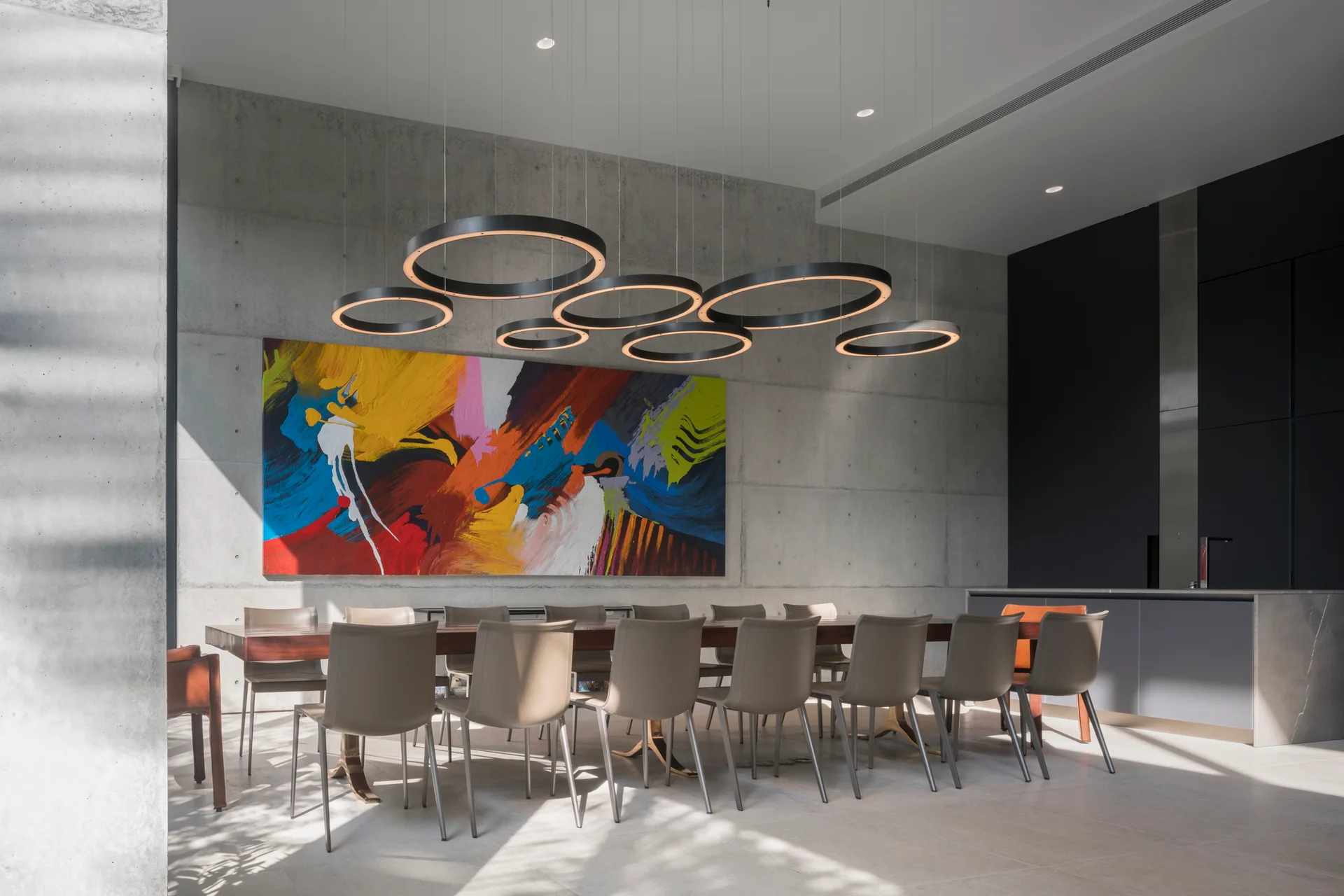 A long wooden dining table and beige chairs sit beneath a cluster of circular ring lights against an exposed concrete wall with colorful art.