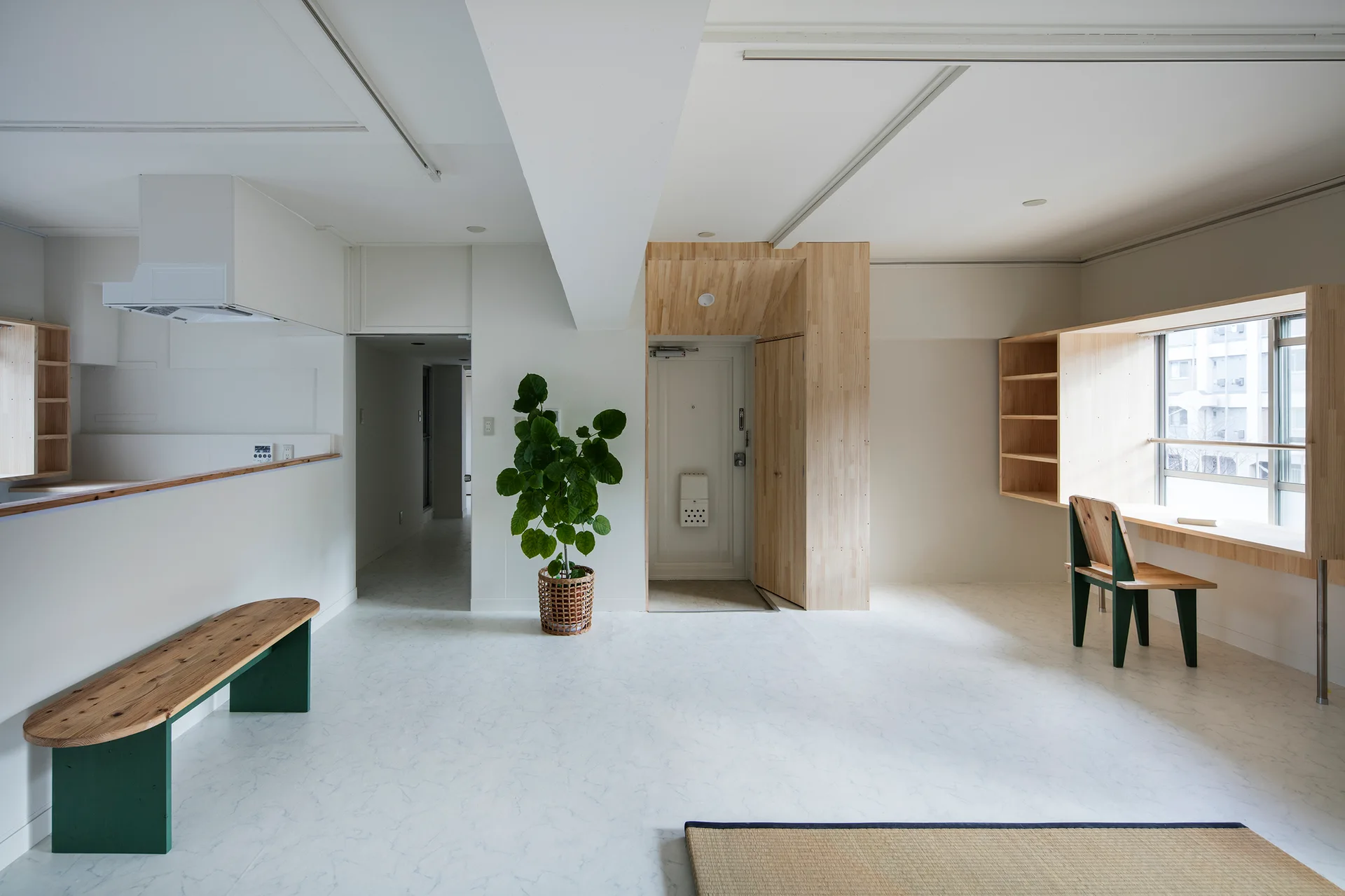 A minimalist white room features a built-in light wood window desk with a green-legged chair, a matching wooden bench, and a tall potted plant.