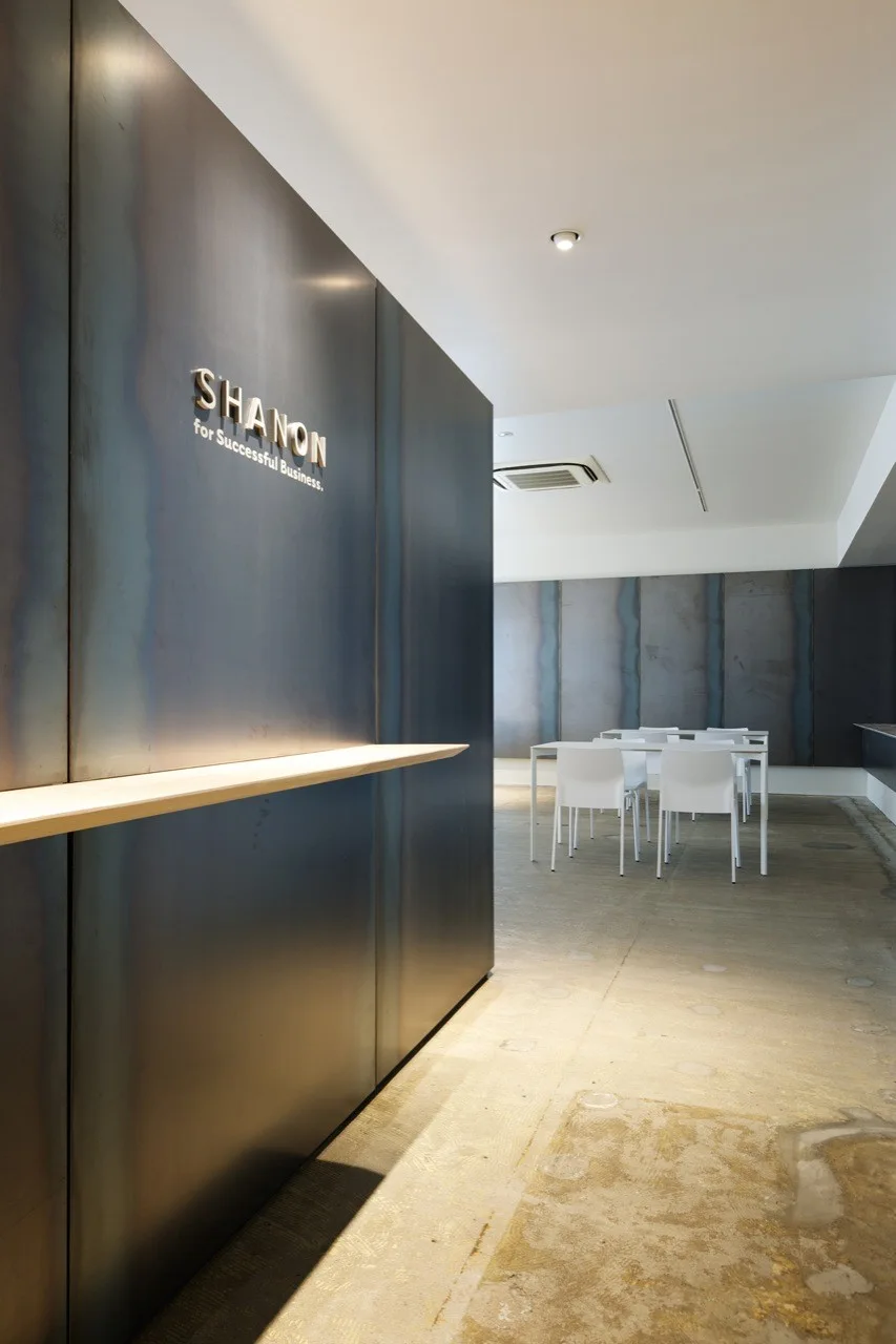 A minimalist office space features a dark metal paneled wall with a light wood ledge, set against polished concrete floors and white chairs.