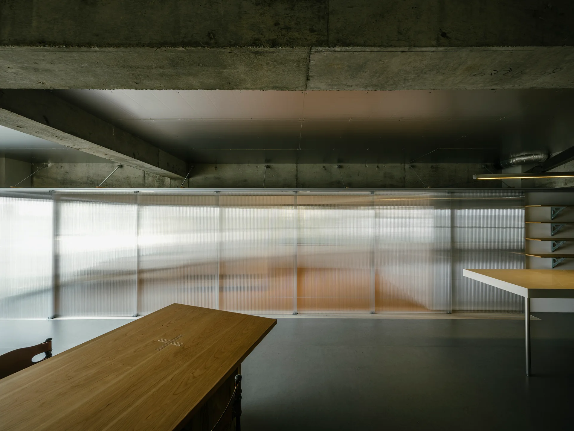 A minimalist interior features a wooden dining table, an exposed concrete ceiling, and a long translucent corrugated sliding screen partition.