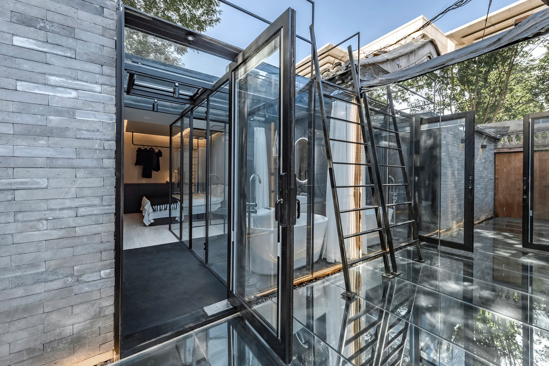 A modern glass-enclosed bedroom and bathroom pavilion features black metal framing, a freestanding white bathtub, glass floors, and grey brick walls.