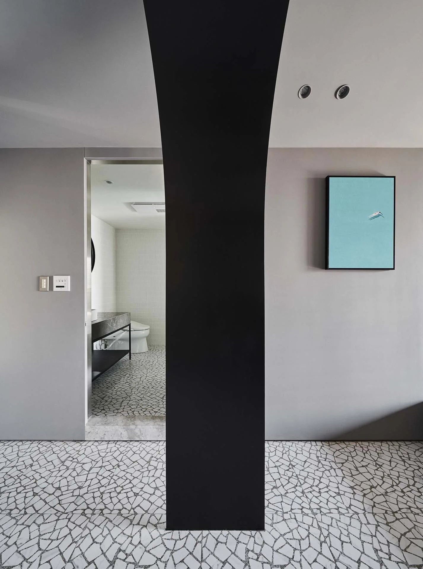 A view through a bold black archway shows a minimalist bathroom with grey walls, a blue framed art piece, and mosaic tile flooring.