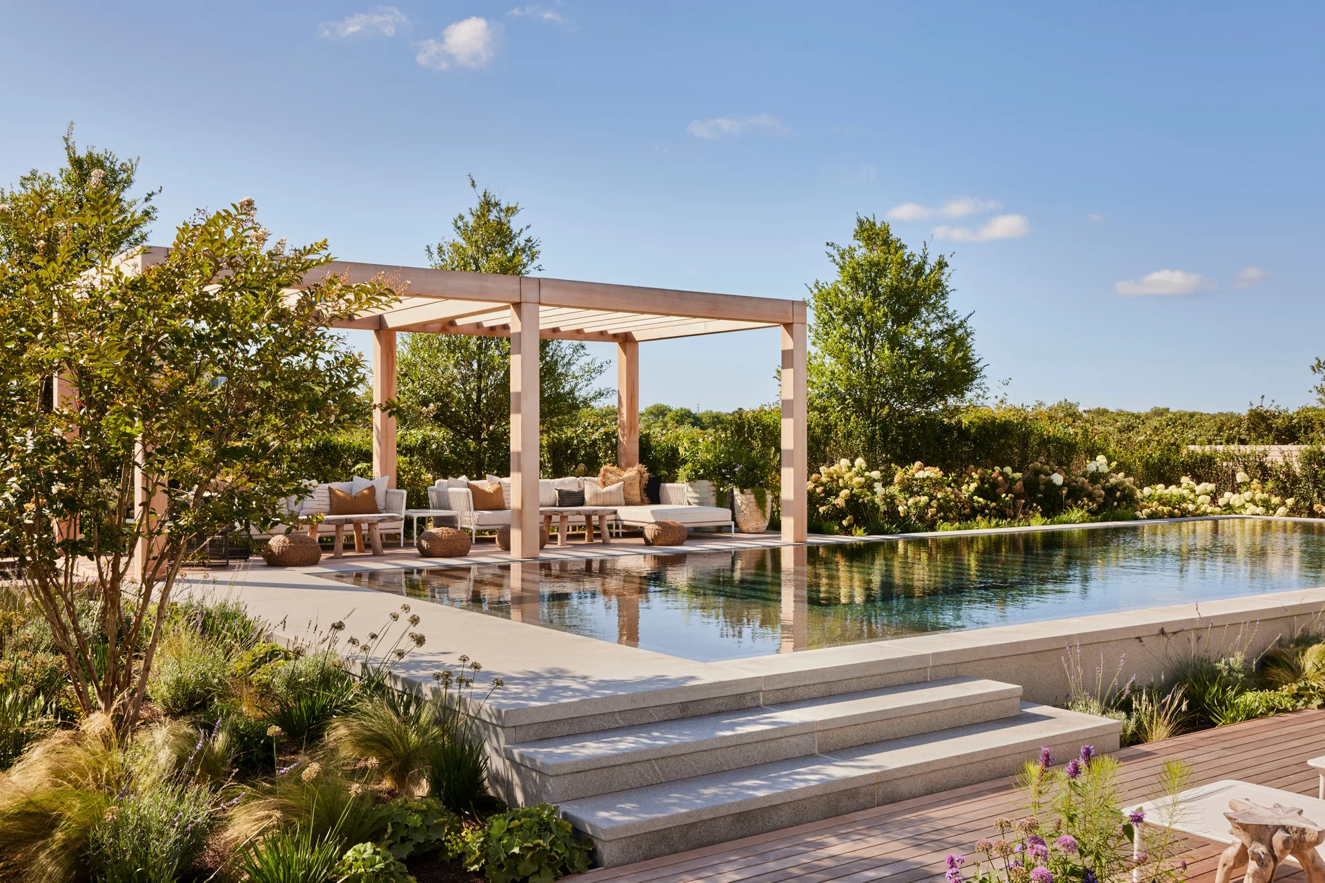 A rectangular backyard swimming pool sits next to a light wood pergola sheltering a beige outdoor sectional sofa, framed by stone steps and green landscaping.