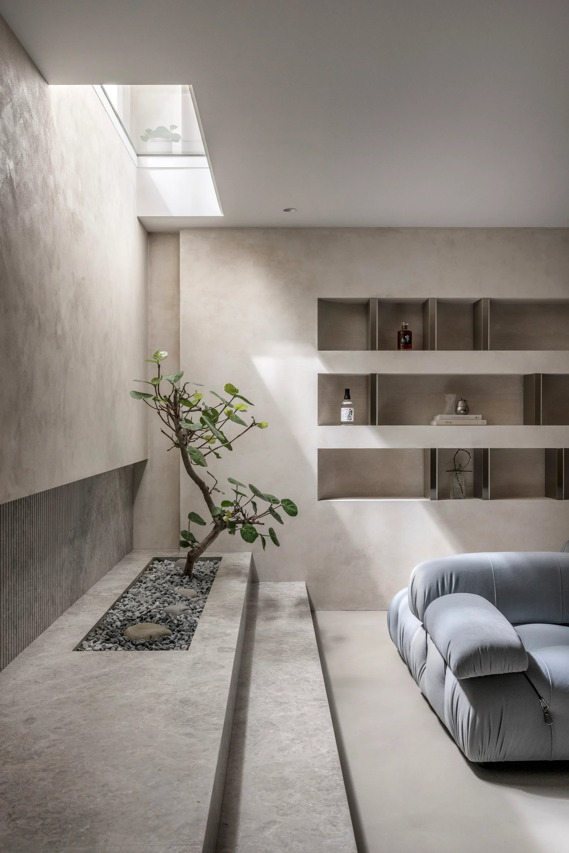 A minimalist living area features a light blue tufted sofa next to a tiered stone indoor planter with a small tree under a skylight.