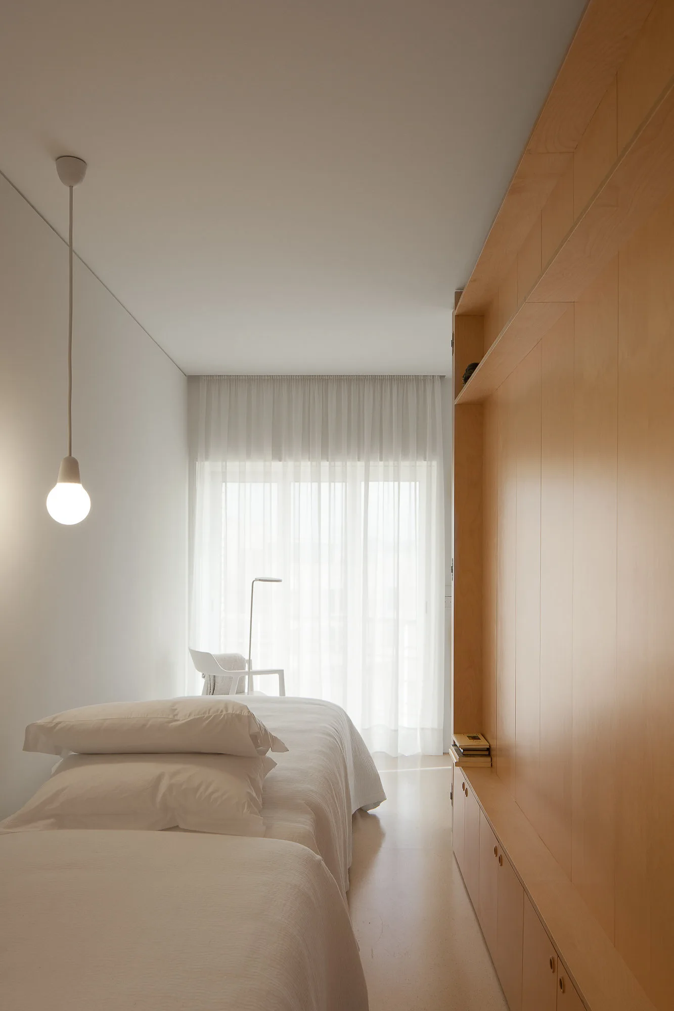 A minimalist bedroom featuring a white bed, a simple pendant light, and a floor-to-ceiling light wood storage cabinet lining the right wall.
