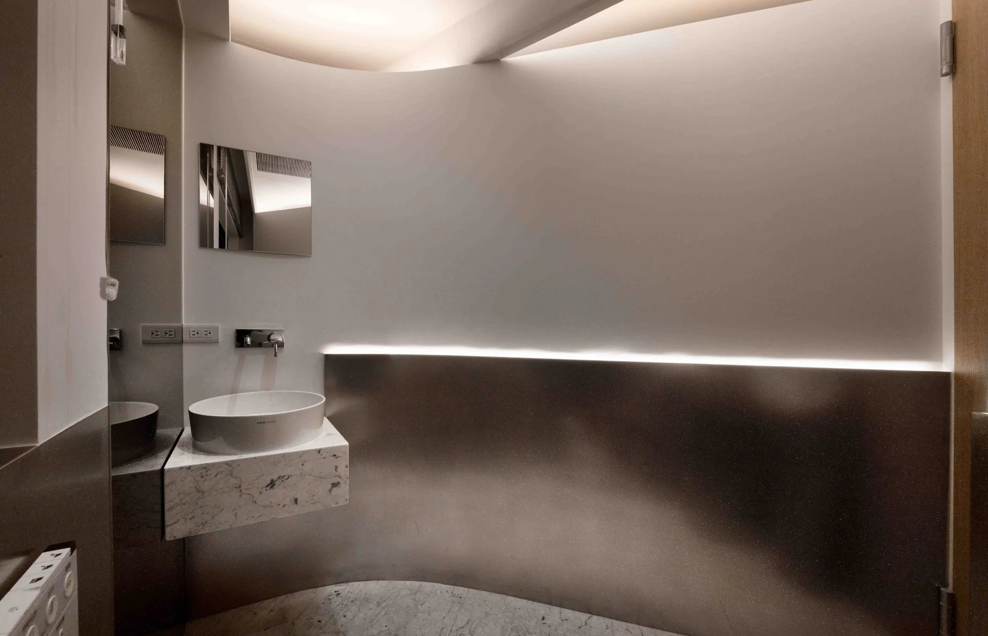 A modern powder room features a floating marble vanity with a white vessel sink against a curved wall with metallic paneling and indirect LED lighting.