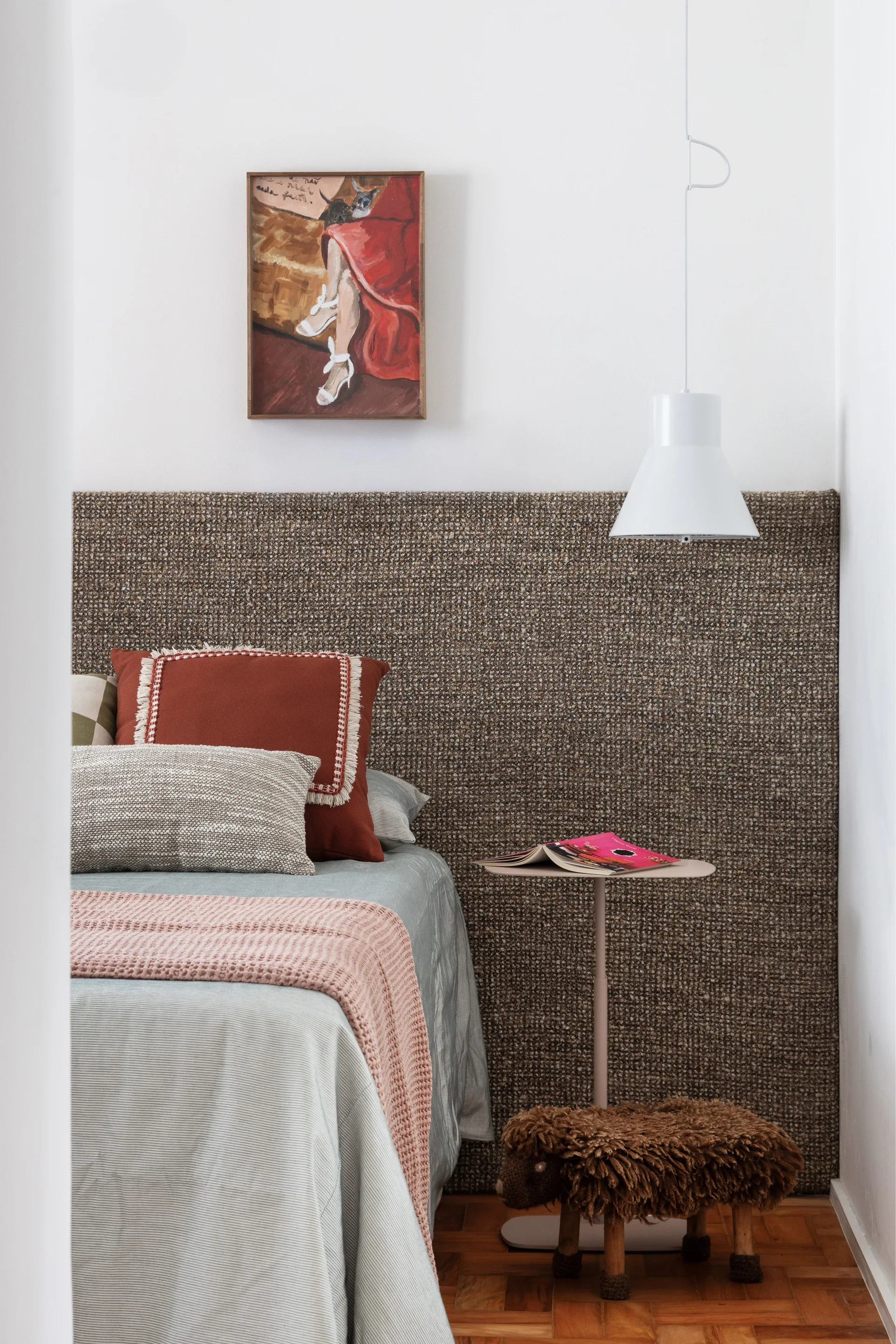 A bedroom features a bed with a tall woven brown headboard, terracotta pillows, a small metal side table, and a hanging white pendant light.