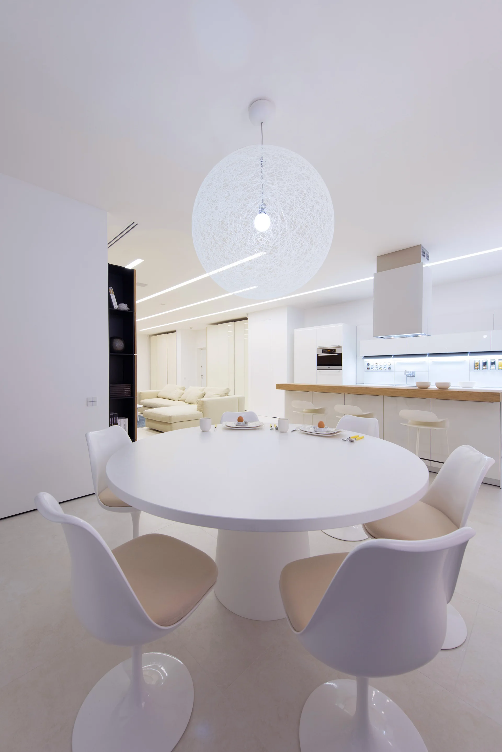 A round white dining table with four molded chairs sits beneath a large spherical string pendant light in a minimalist, open-concept home.
