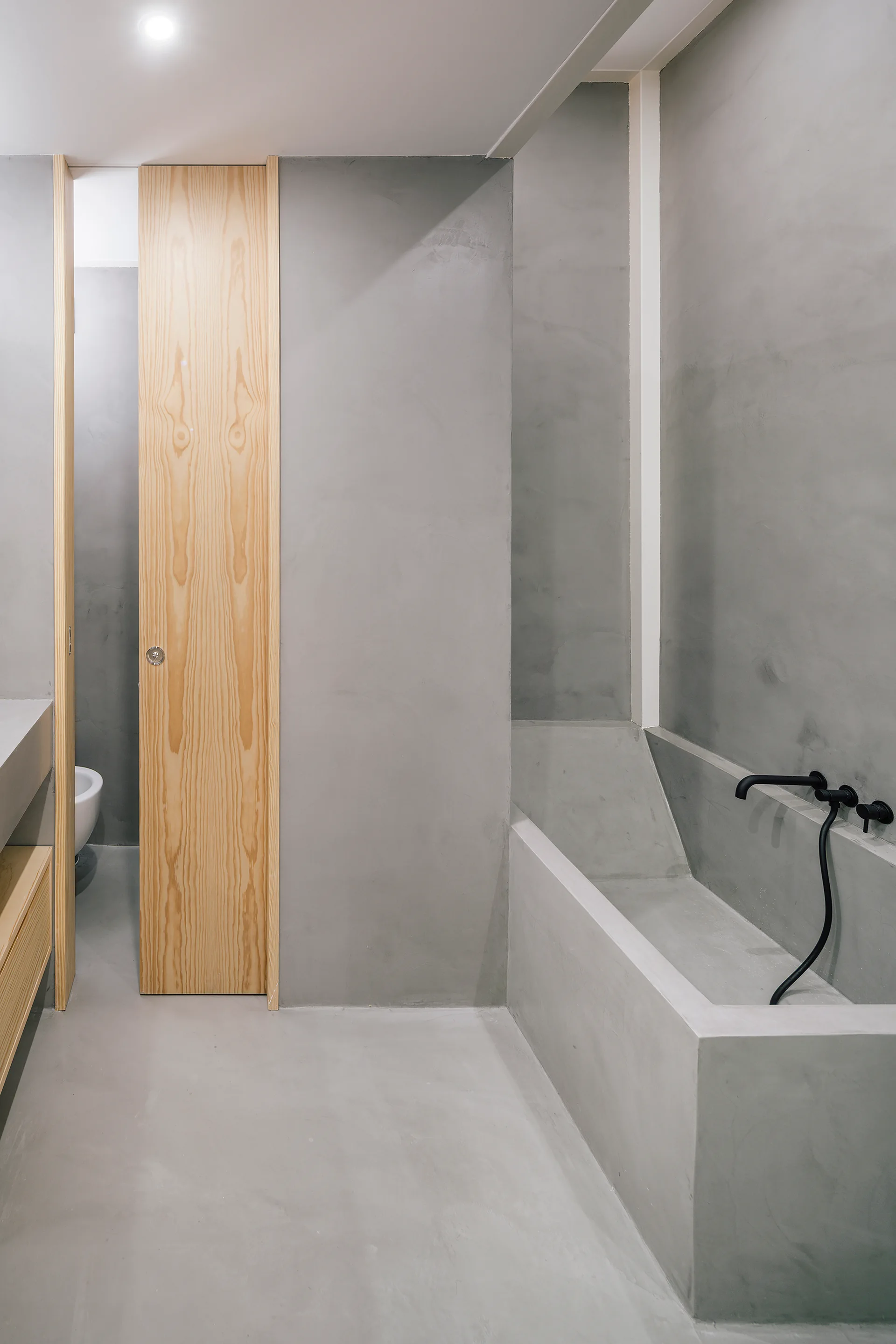 A minimalist grey bathroom featuring a built-in concrete bathtub, a tall light wood door, and matte black wall-mounted fixtures.
