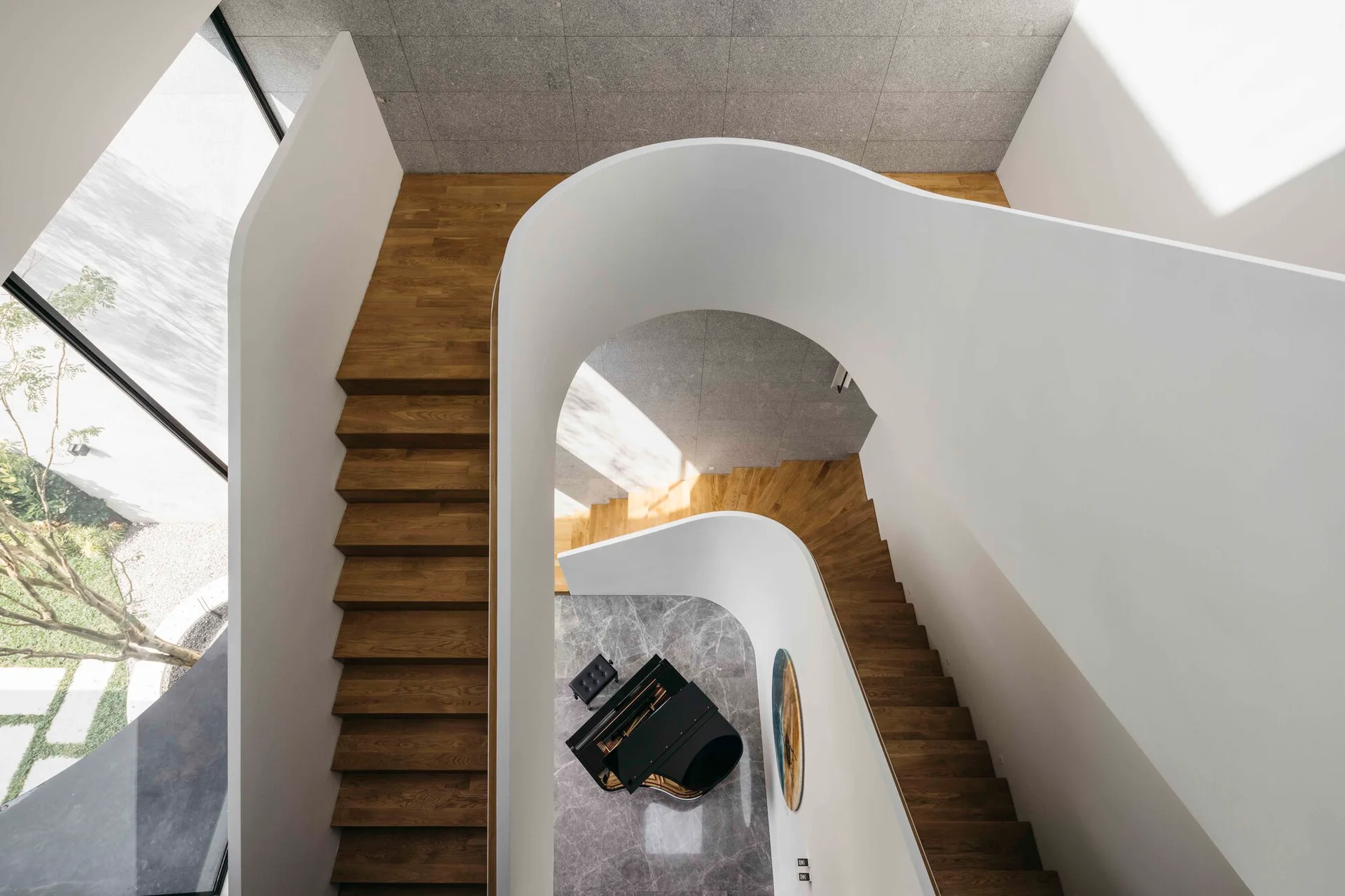 A high-angle view of a sculptural curved white staircase with wooden treads winding down toward a black grand piano on a polished grey stone floor.