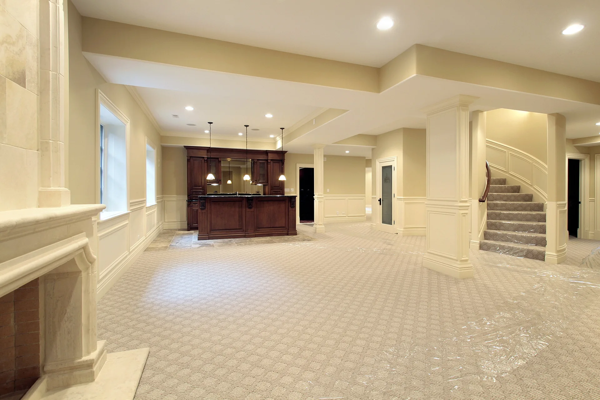 A spacious basement lounge features beige walls with classic wainscoting, a carpeted floor, a stone fireplace, and a dark wood wet bar in the background.