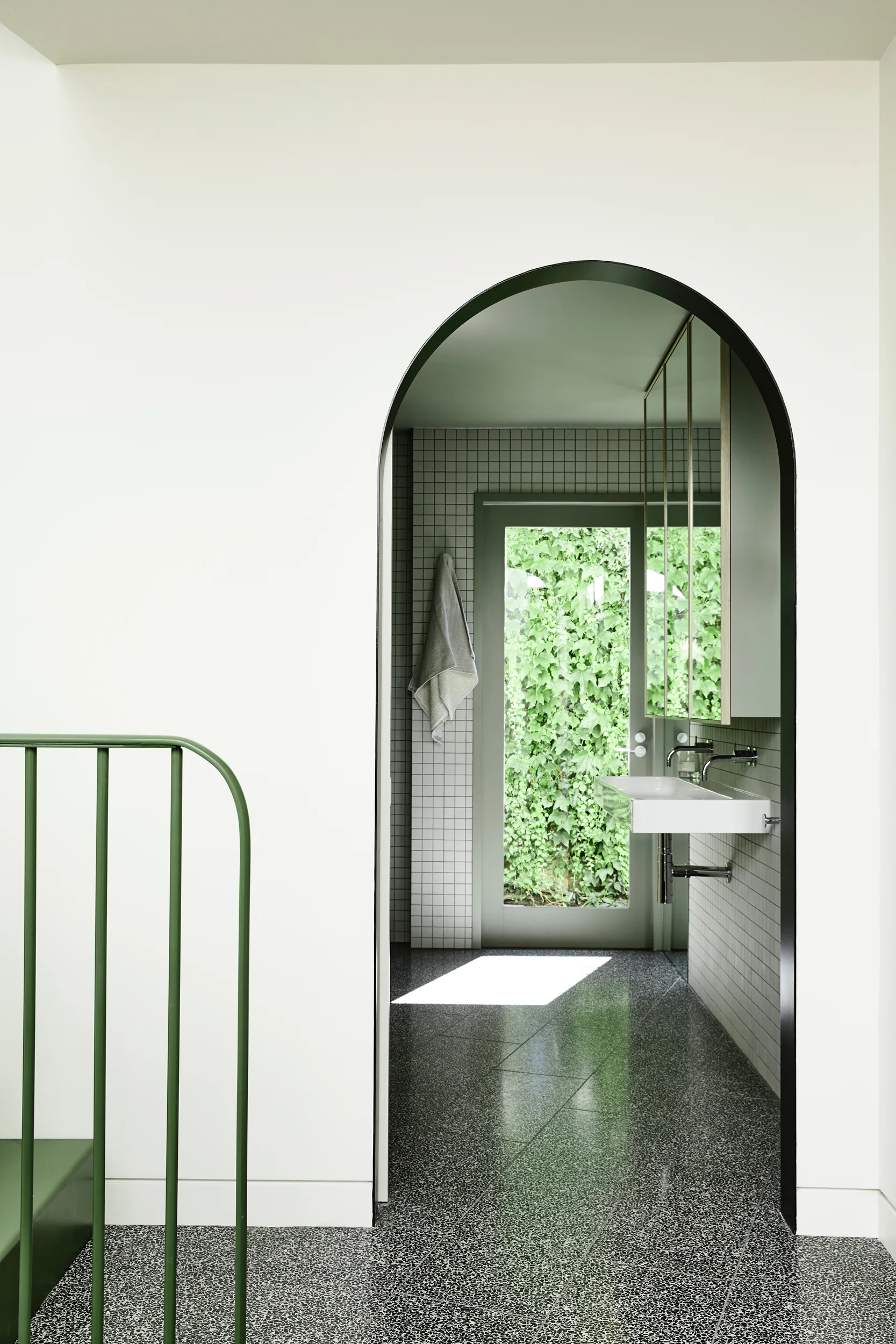 A black-painted arched entryway opens into a bathroom featuring a wall-mounted sink, small square white wall tiles, and dark terrazzo flooring.