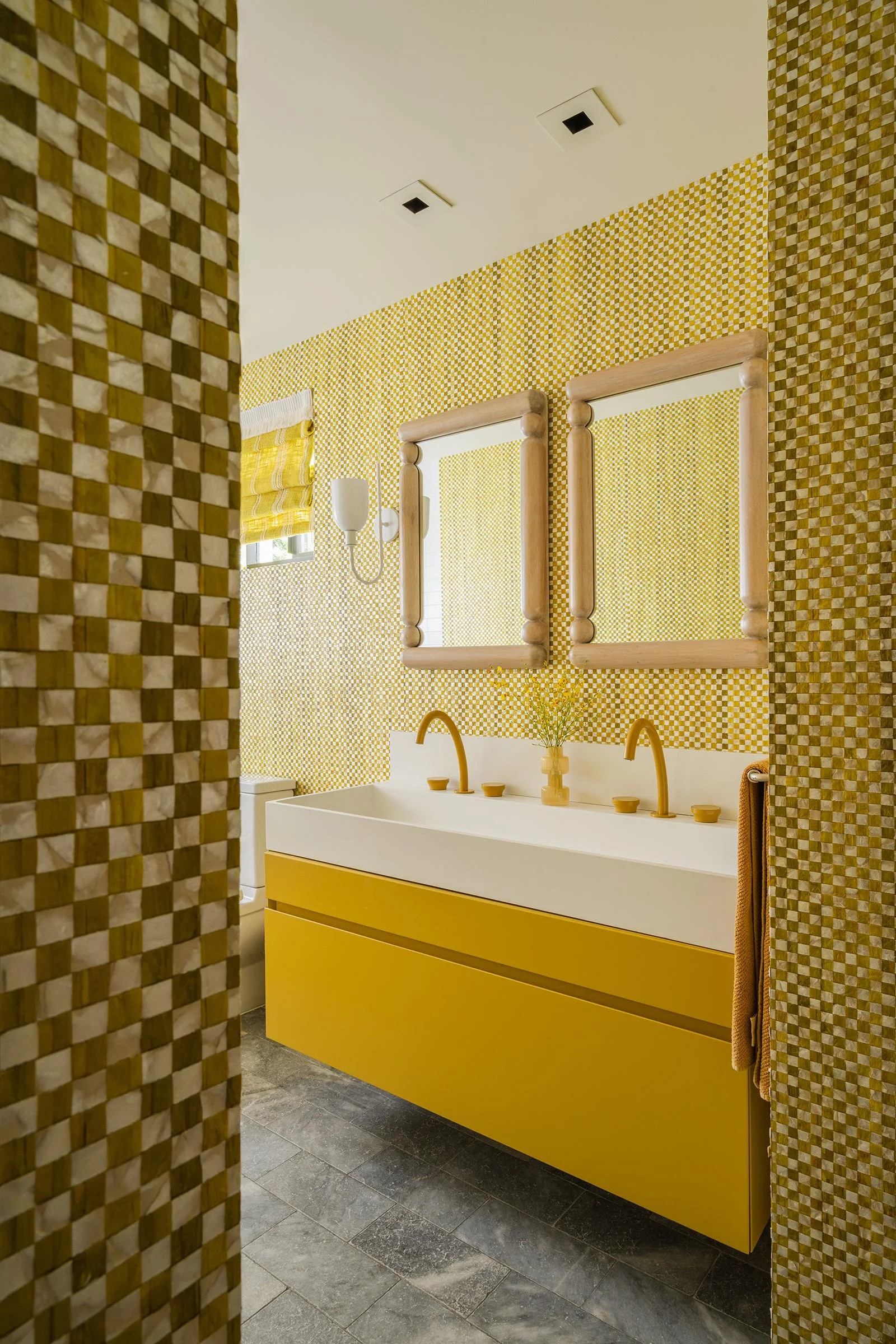 A modern bathroom features yellow and white checkered wallpaper, two rectangular wooden mirrors, and a yellow floating vanity with a white trough sink.