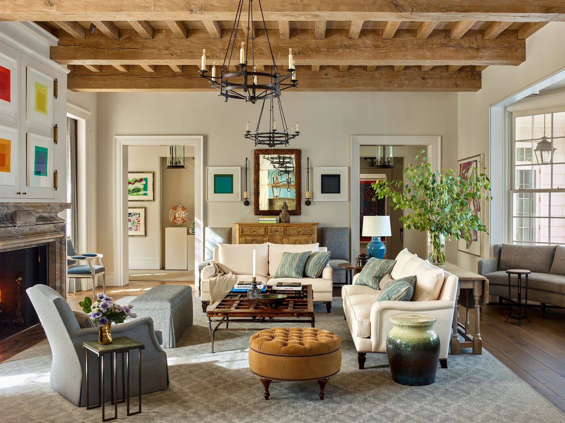 A spacious living room featuring exposed wood ceiling beams, cream sofas, a round tufted ottoman, and black iron chandeliers.