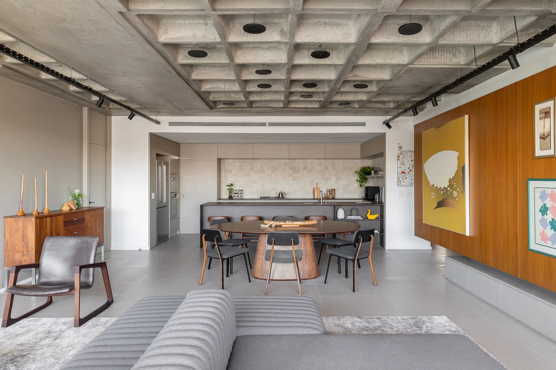An open-plan room features an exposed concrete waffle ceiling, an oval wood dining table with grey chairs, and a grey sofa.