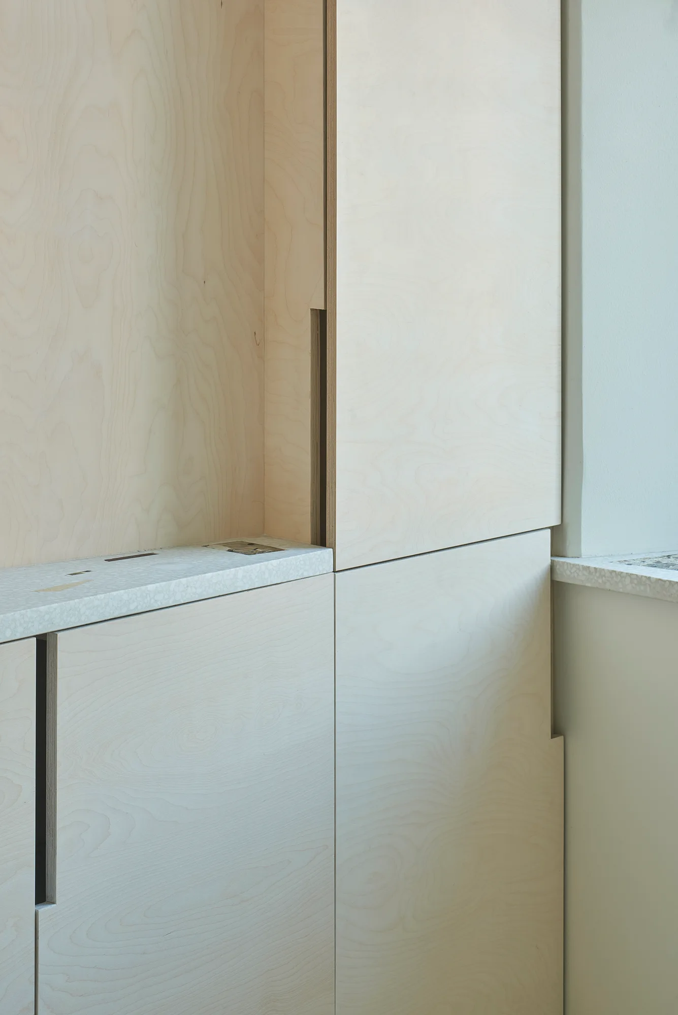Close-up of minimalist plywood cabinetry with integrated vertical handles and a speckled terrazzo countertop next to an off-white wall.